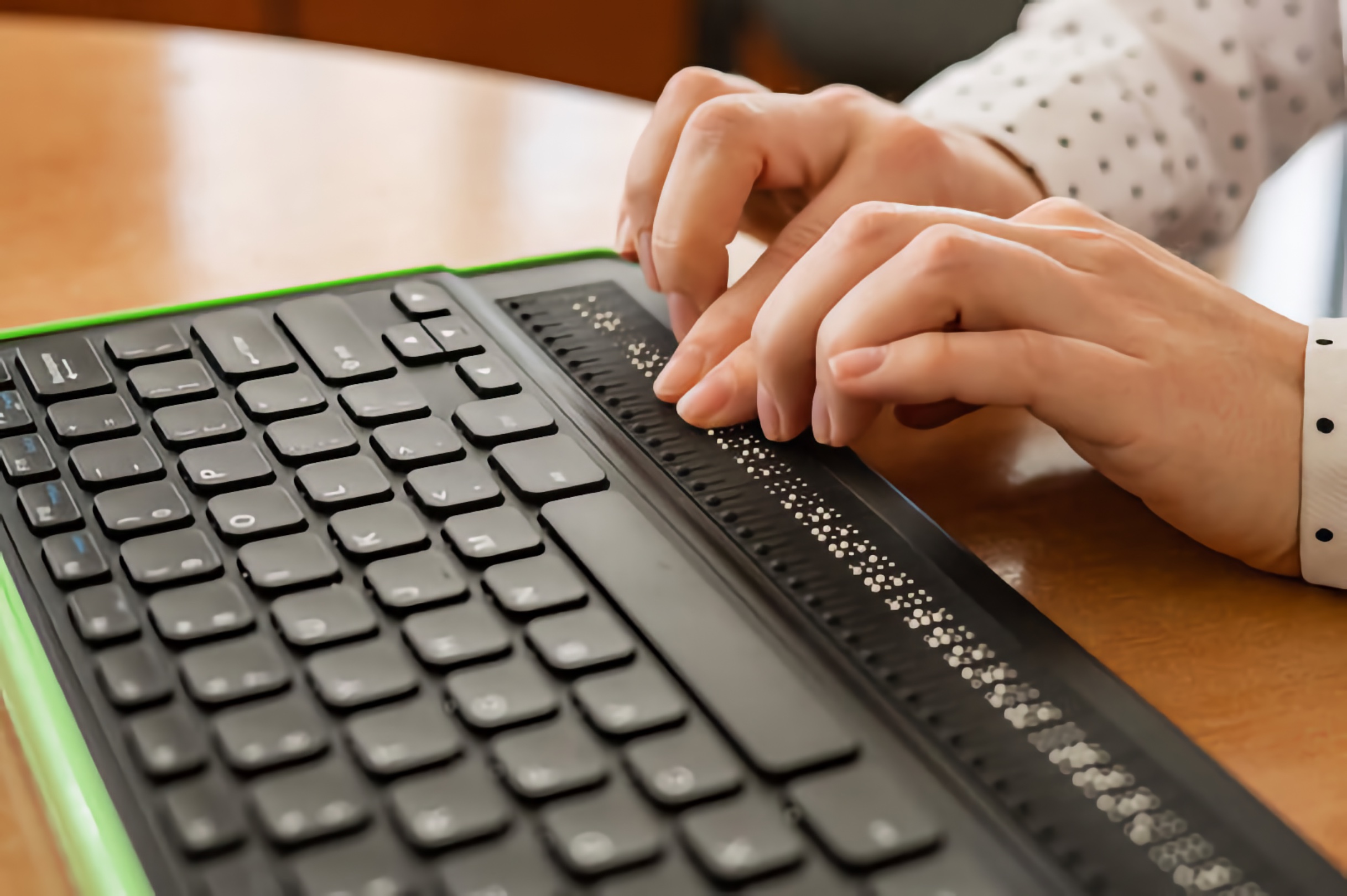 Close-up of hands reading a refreshable braille display beside a computer keyboard on a wooden table