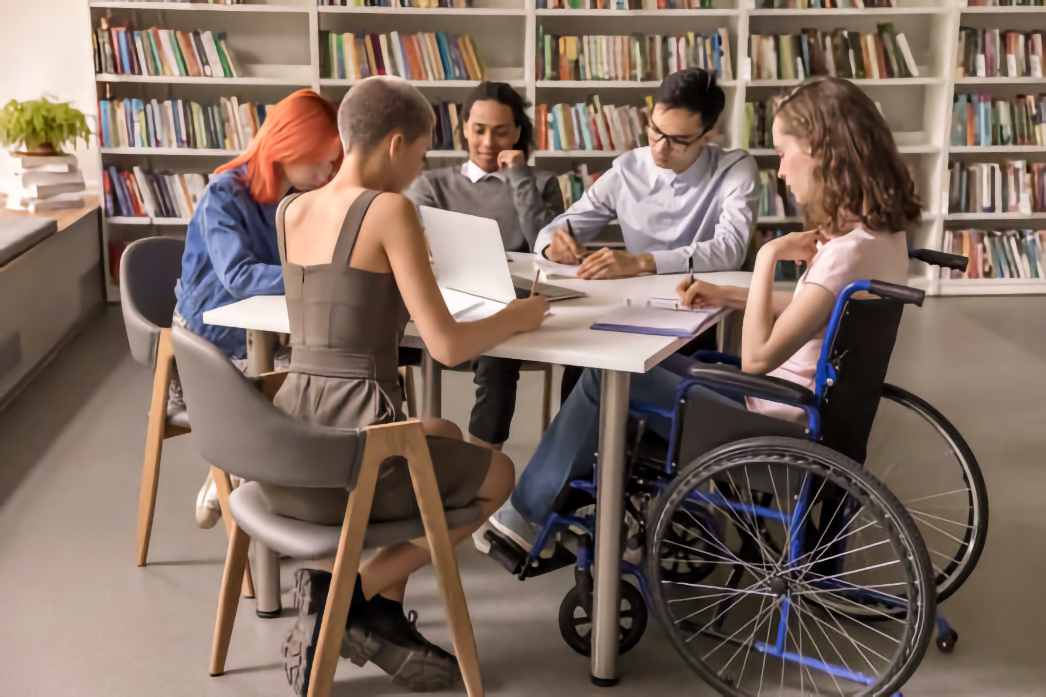 Five multiracial young adults, including one wheelchair user, study together at a library table.