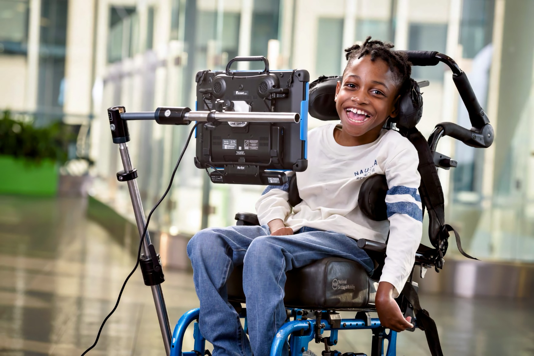 A Black boy smiles broadly from his wheelchair beside a speech device mounted on an adjustable arm at his eye level in a glass atrium.