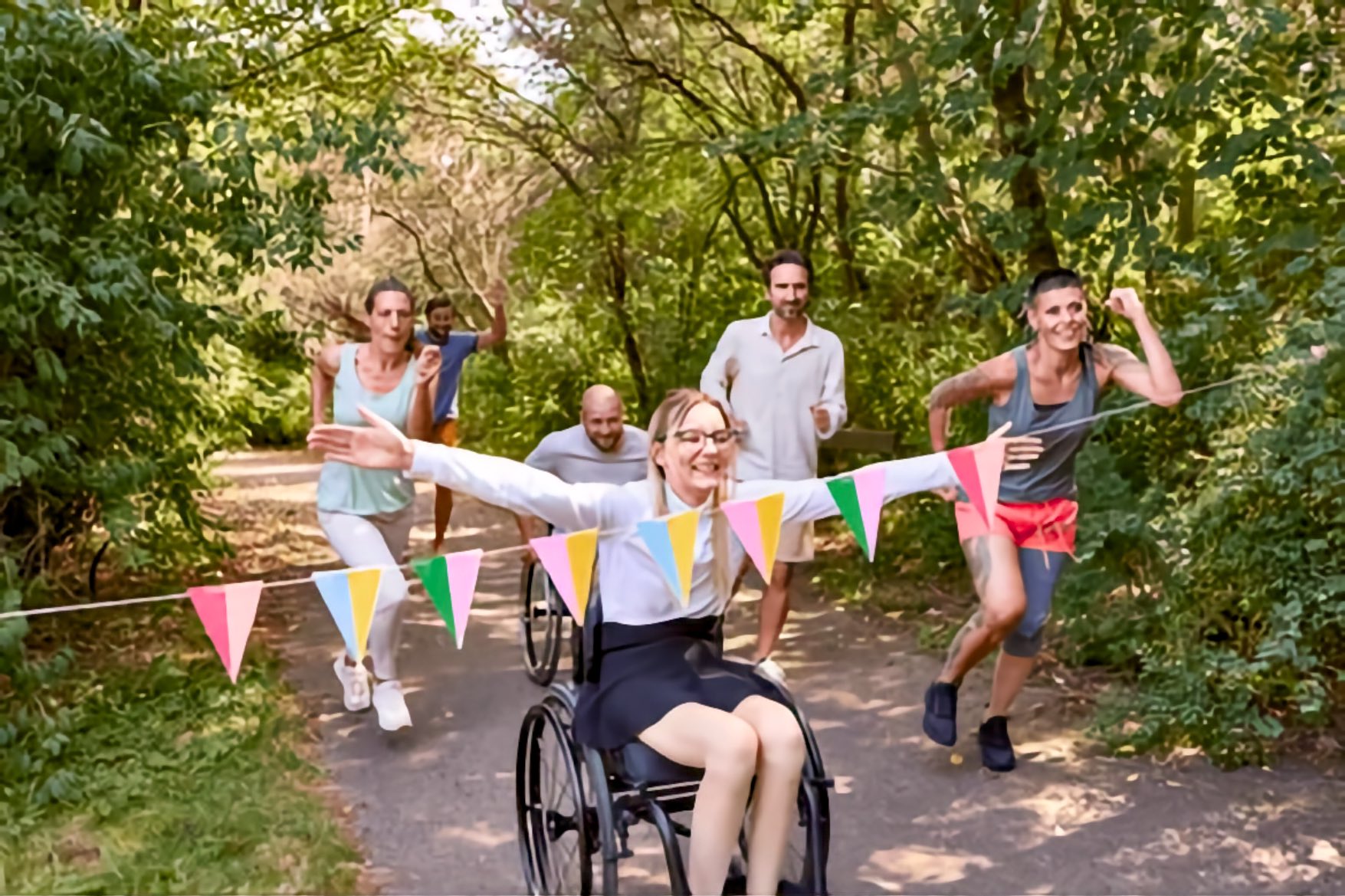 A white woman in a wheelchair crosses a bunting finish line on a wooded path, arms spread wide, as Black and white runners cheer behind her.