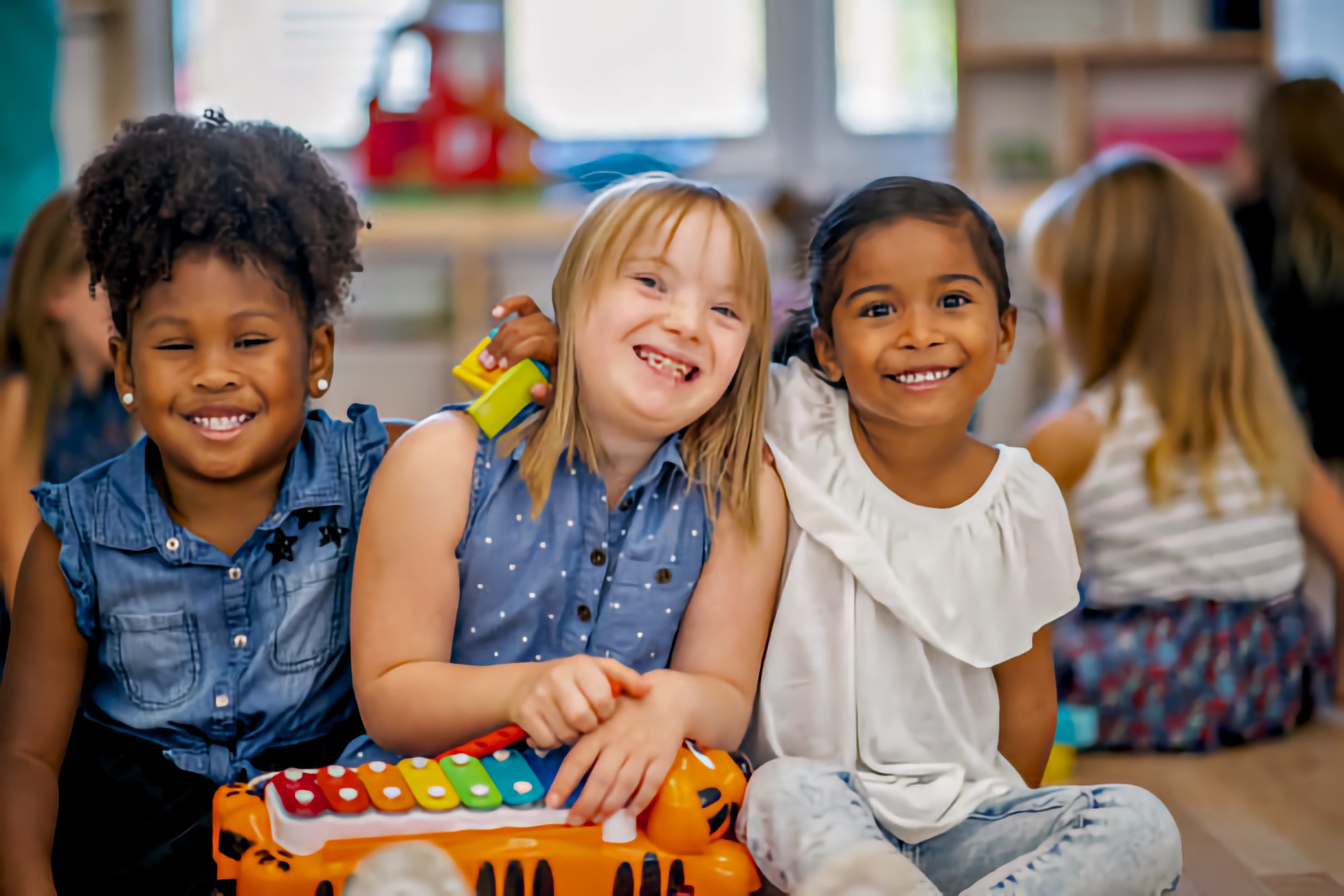 Three girls of different races sit arm in arm on a classroom floor behind a toy xylophone, smiling at the camera as other children play behind them.