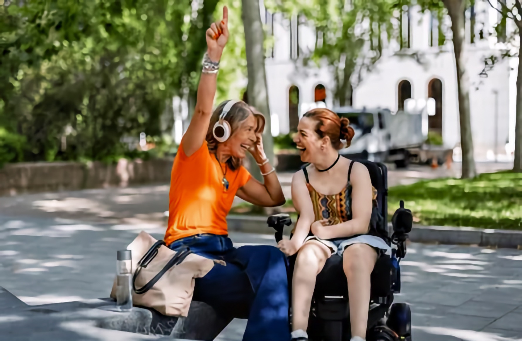 Two women laugh together on a low stone ledge in a plaza, one wearing headphones with a hand raised, the other seated in her power wheelchair.