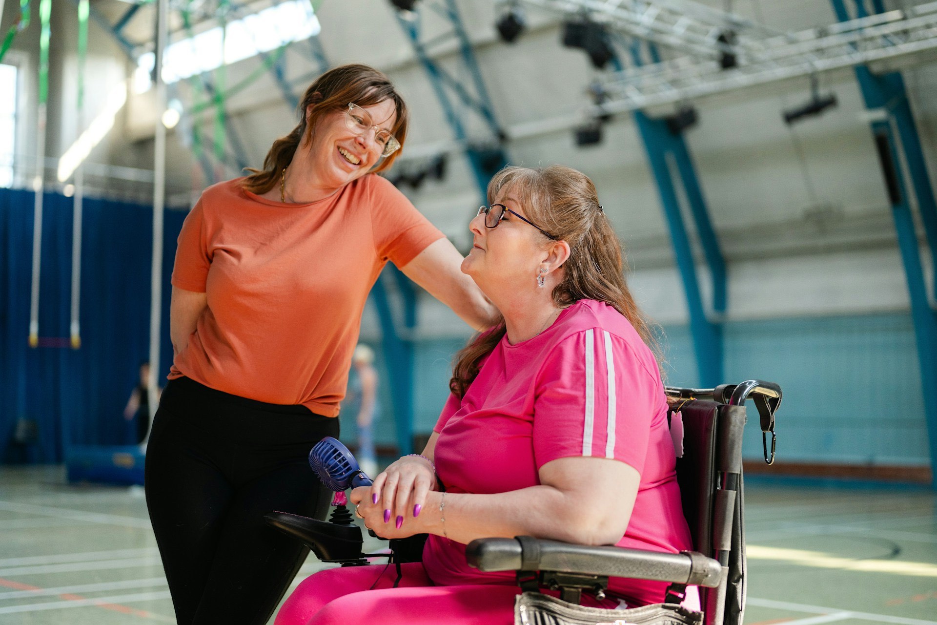 Two women talk closely in an indoor sports hall with court lines on the floor, gymnastics rings hanging on the left and a curtain across the back wall. One woman sits in a power wheelchair with her right hand resting on the joystick control and a small handheld fan in her left hand, looking up and smiling. The other woman leans toward her with one hand on the back of the wheelchair, laughing with her.