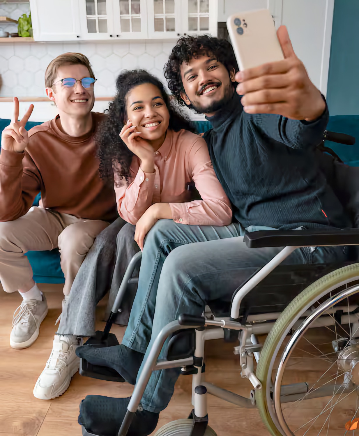 Three young adults pose for a selfie in a kitchen. A South Asian man using a manual wheelchair holds the phone out and smiles. A Black woman leans on his shoulder and a white man beside her raises a peace sign.
