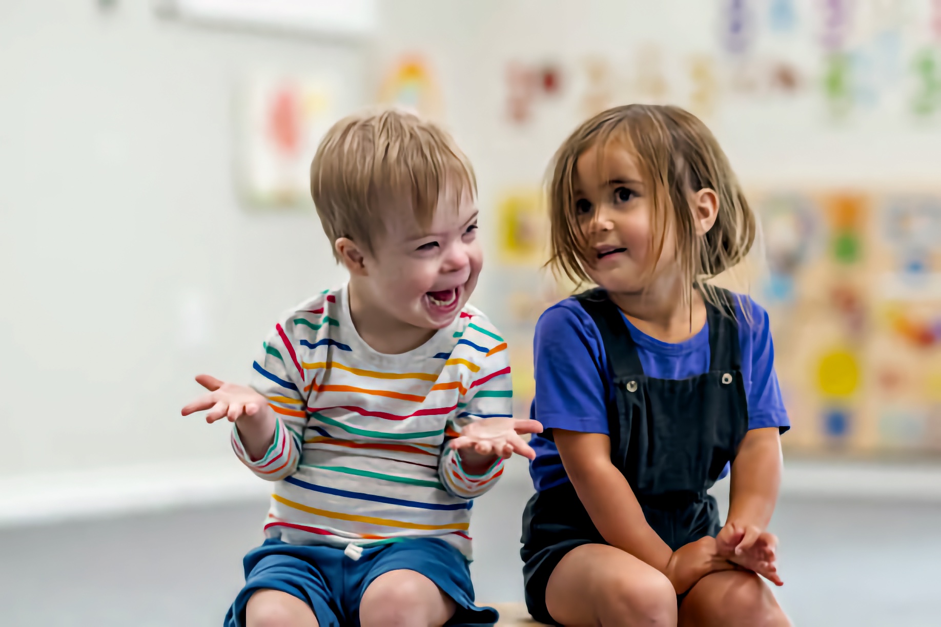 A boy with a disability laughs with his palms raised as a girl beside him smiles toward him. Sitting together in a classroom, they share a moment of connection and belonging.