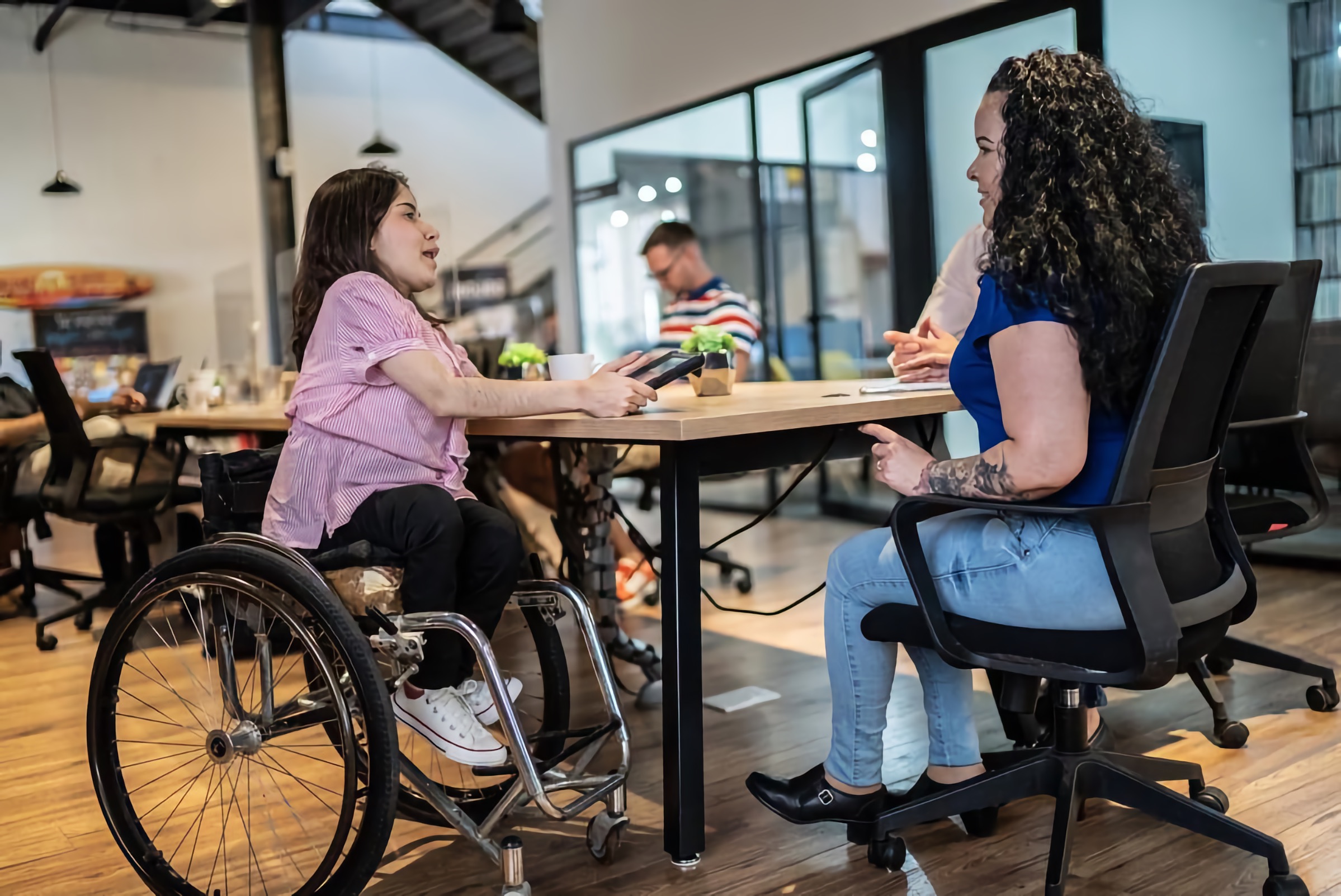 Two colleagues meet at a shared desk; one uses a wheelchair and holds a tablet in a modern office.