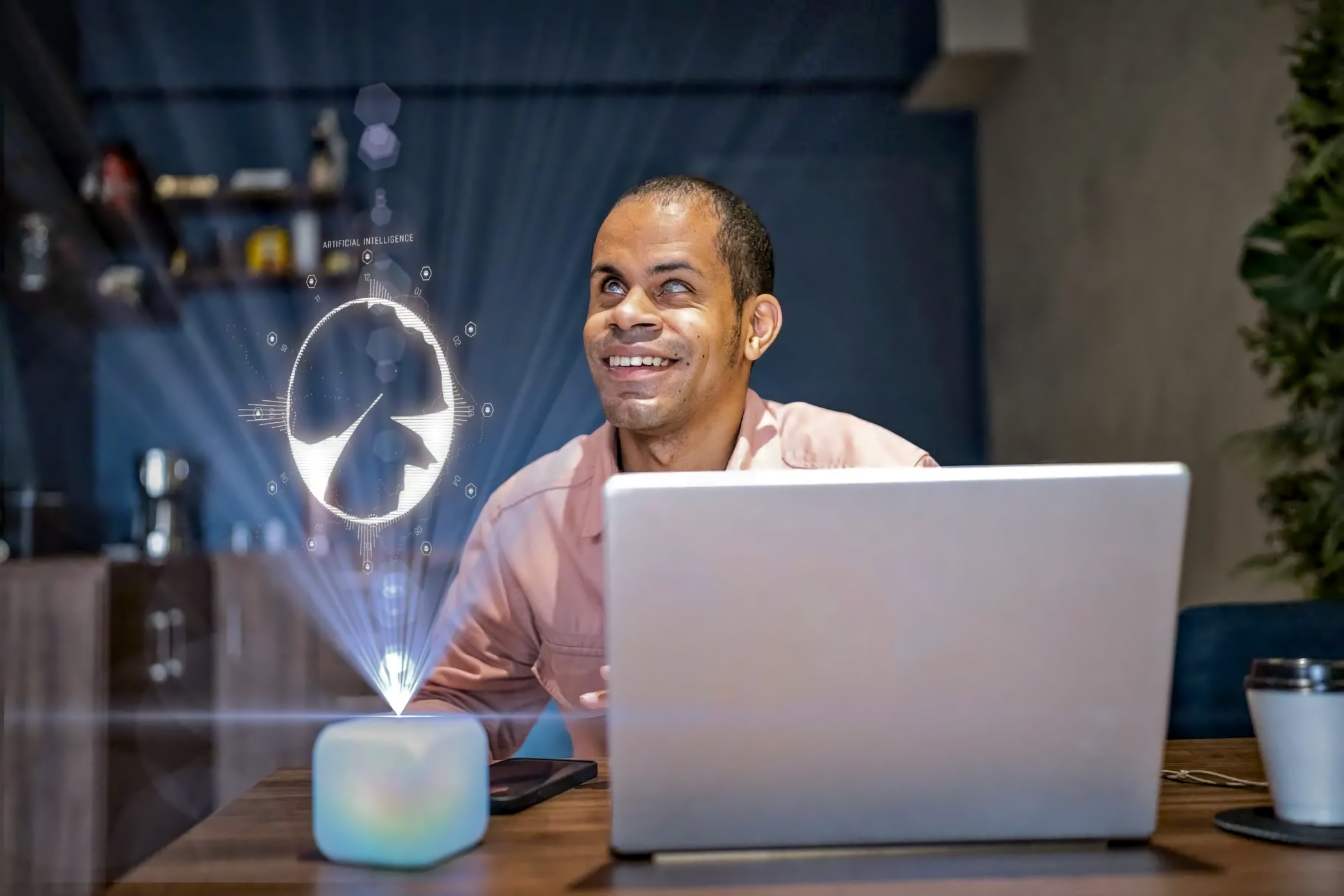 A smiling Blind Black man in his 30s sits at a desk with his head tilted upward in the direction of a glowing holographic artificial intelligence projection emanating from a small illuminated device, with an open laptop on the desk in front of him.