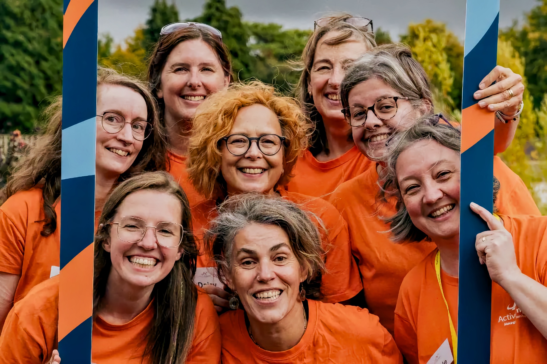 Eight adults in orange T-shirts smiling outdoors and holding a blue-and-orange prop frame; trees and clouds behind.