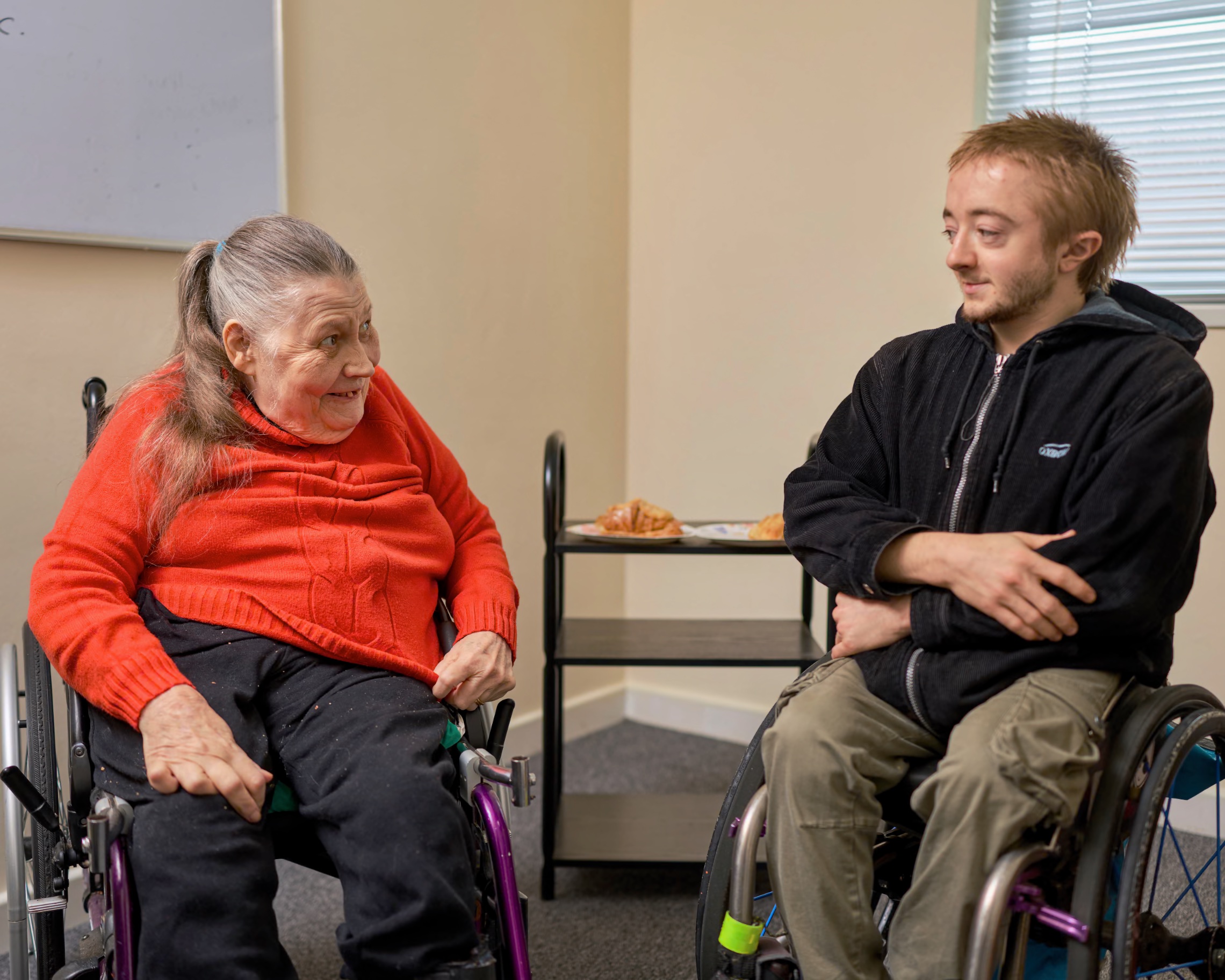 Elder adult woman (81) and young adult man (25), both manual wheelchair users, sit indoors facing each other in conversation; their wheelchairs’ push-rims and footplates are visible; behind them are a whiteboard, window blinds and a small trolley with food