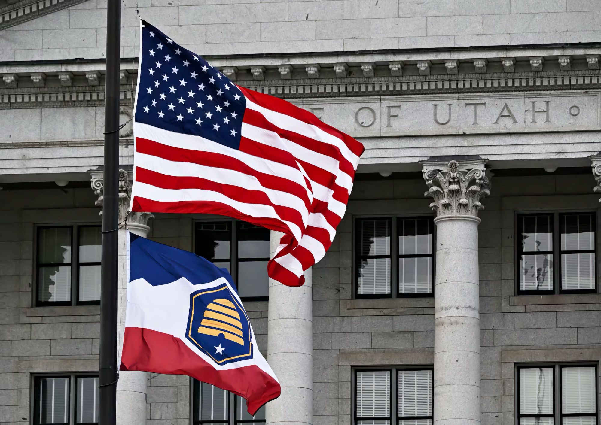 Two flags on tall flagpoles whip beneath a cloudy sky outside the Utah State Capitol