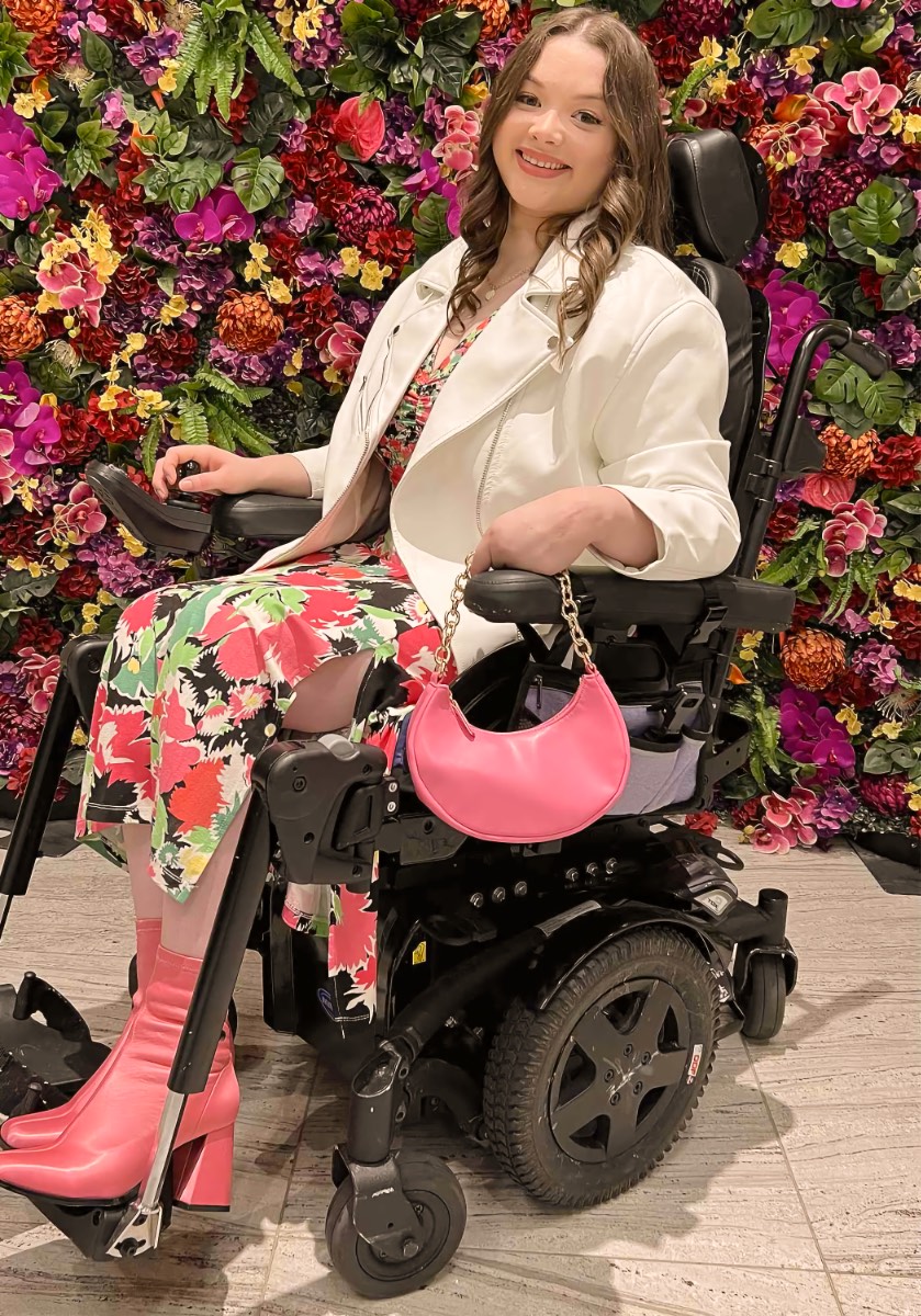 A smiling adult woman with long wavy brown hair sits in a black power wheelchair with a headrest, footplates, and a joystick control beneath her hand, in front of a dense wall of artificial flowers in pink, purple, red, orange and yellow with green foliage.