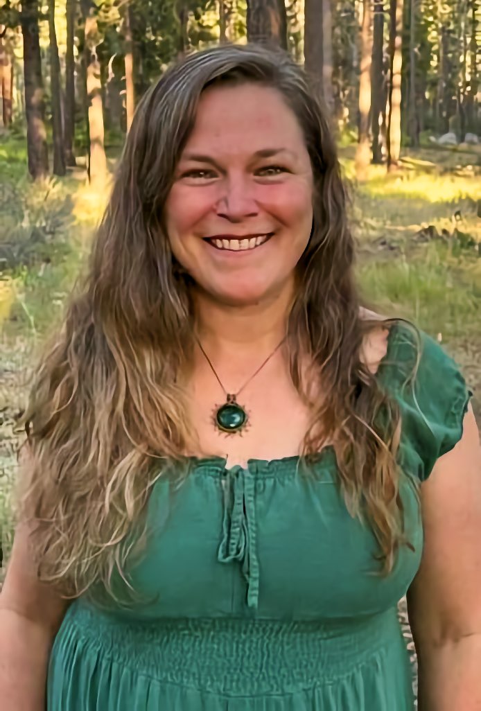 A woman with long wavy hair smiles directly at the camera, standing outdoors in an evergreen forest with low sunlight coming through the trunks behind her.