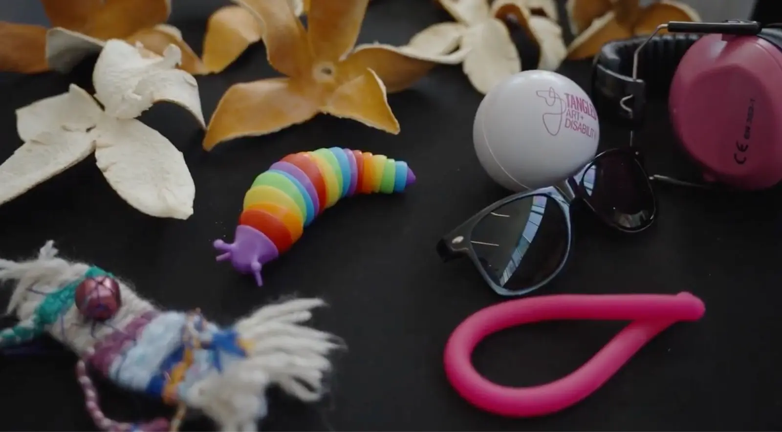 Dried orange peels with sensory tools—rainbow fidget slug, pink loop, sunglasses, and over-ear defenders—on a black surface.