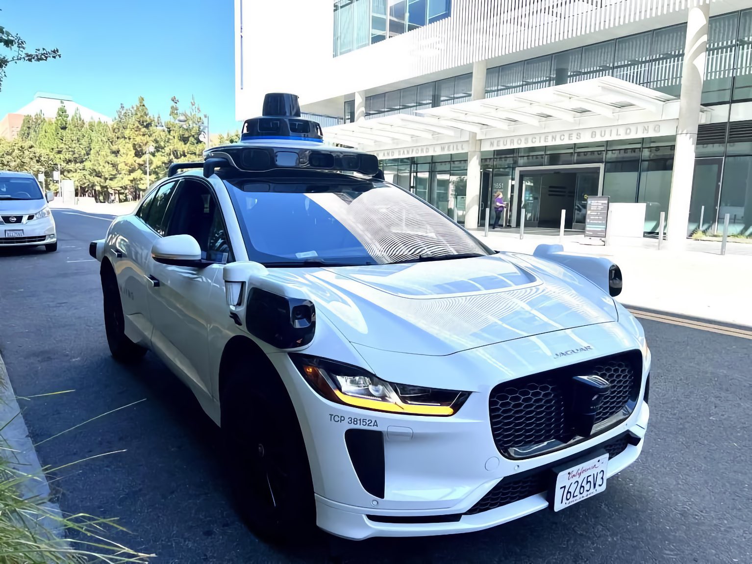 White Jaguar autonomous test car with roof lidar and body-mounted sensors stopped outside a Neurosciences Building entrance on a step-free city street under a clear sky.