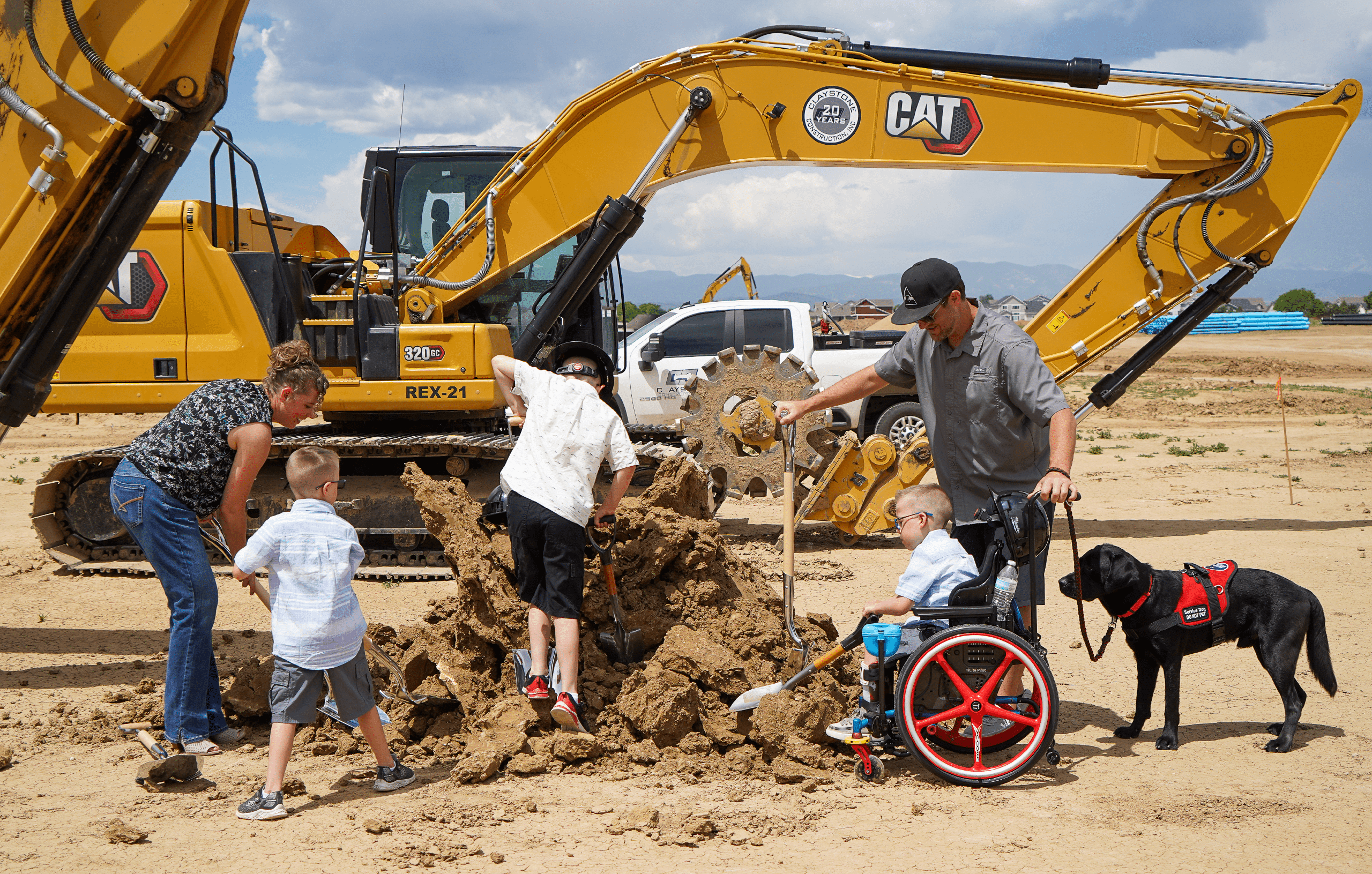 A boy using a wheelchair digs into a mound of dirt with a shovel while his service dog, a black Labrador with a red vest, stands nearby. Two other children, a man, and a woman also dig with shovels. In the background, a large yellow CAT excavator is visible with its arm raised, ready for construction.