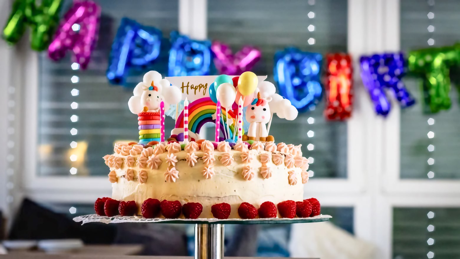 A frosted layer cake ringed with raspberries and topped with unicorn figures, a rainbow, and candles, set before foil balloons spelling Happy Birthday.