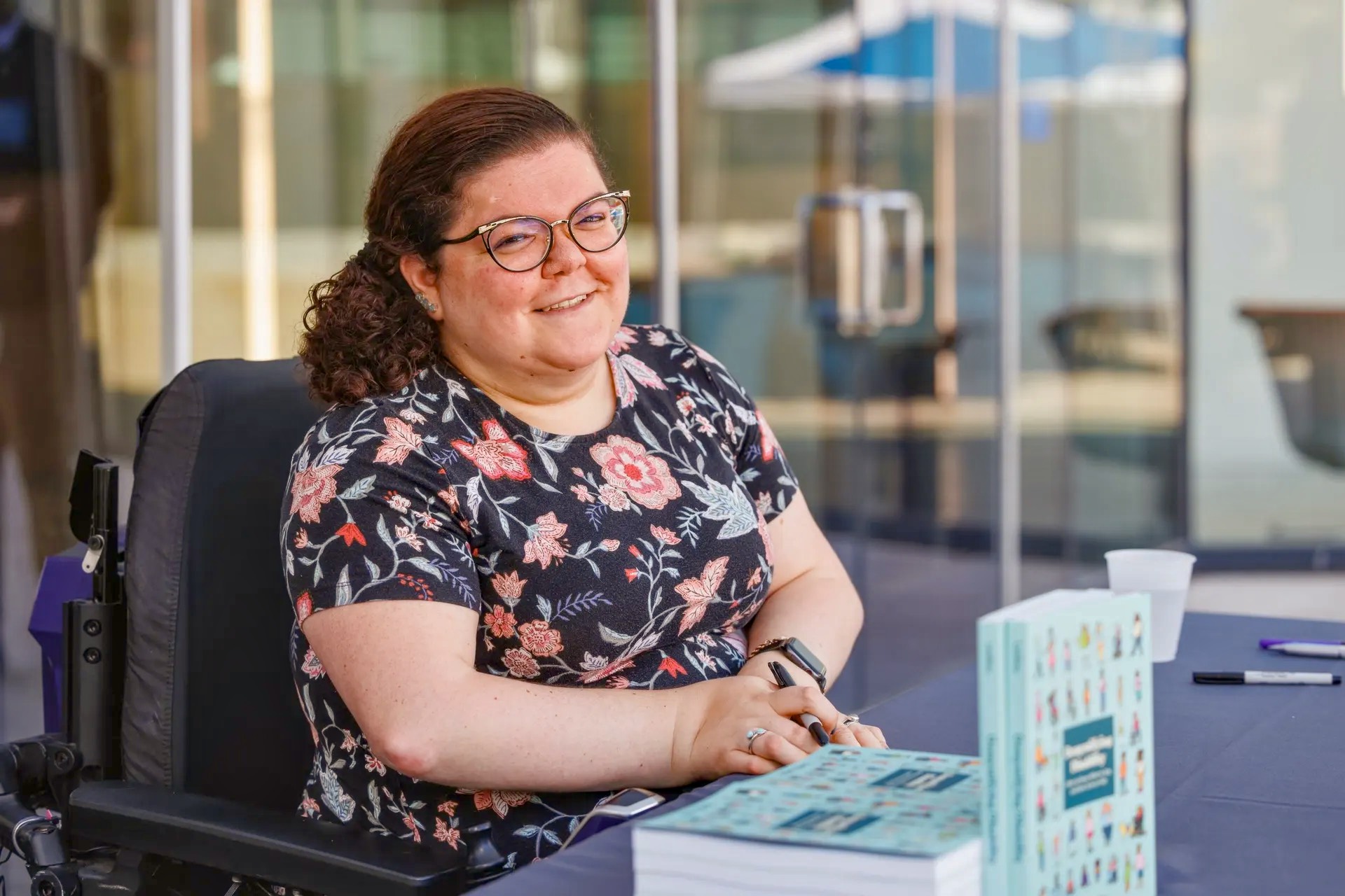 A smiling woman in a wheelchair sits at a table, pen in hand, beside stacked copies of a book.