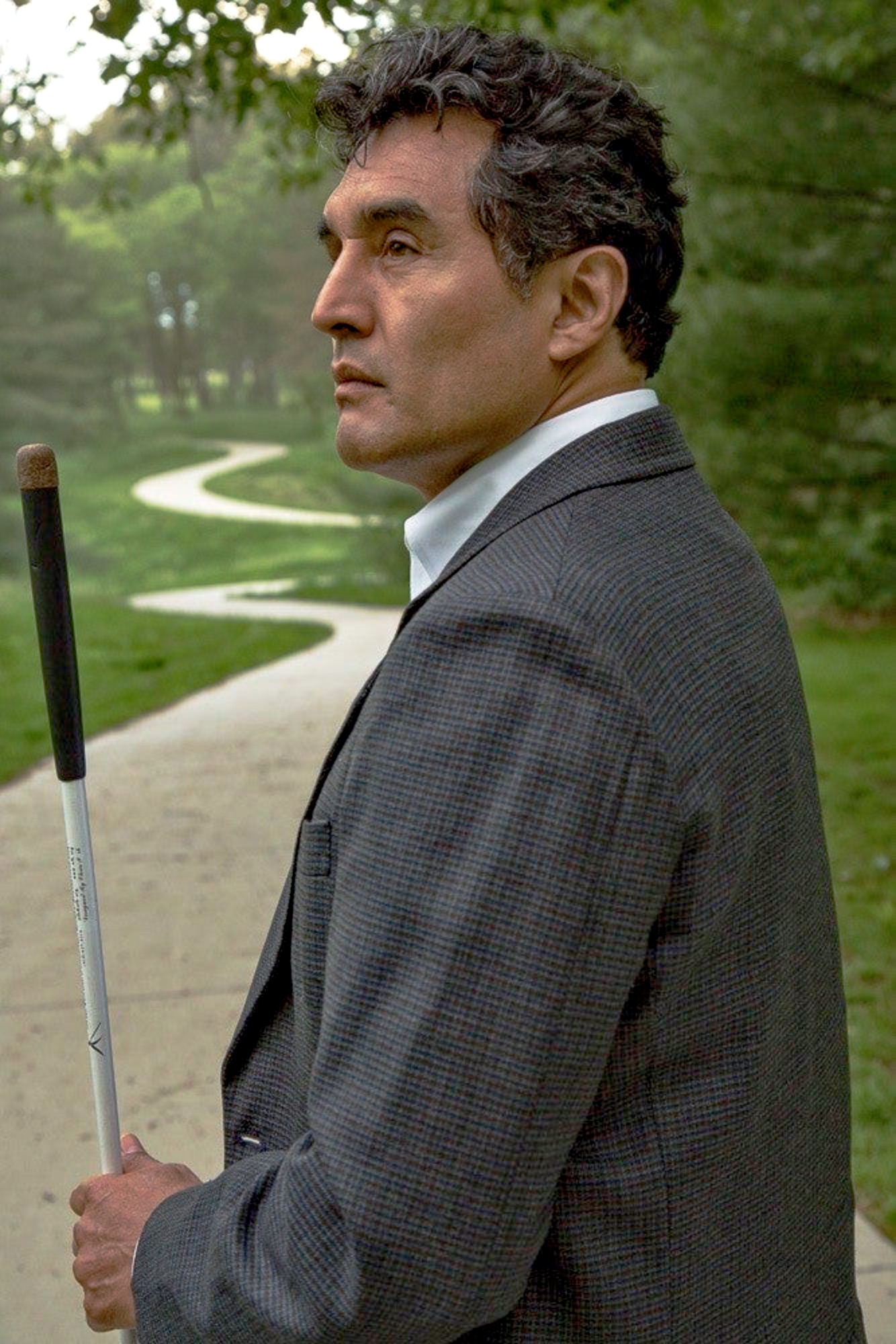 Blind man with wavy dark hair, stands on a paved path in a green park, holding a long white cane. He wears a dark checkered blazer over a white shirt and has a serious, focused expression.