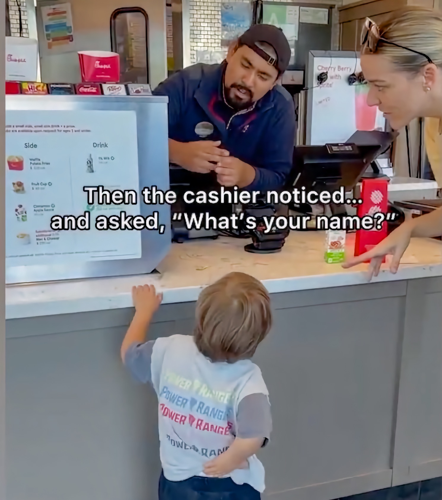 At a fast-food counter, a bearded male cashier in a cap leans forward speaking with a small boy in a Power Rangers T-shirt while an adult woman stands nearby; on-screen text reads, “Then the cashier noticed… and asked, ‘What’s your name?’”