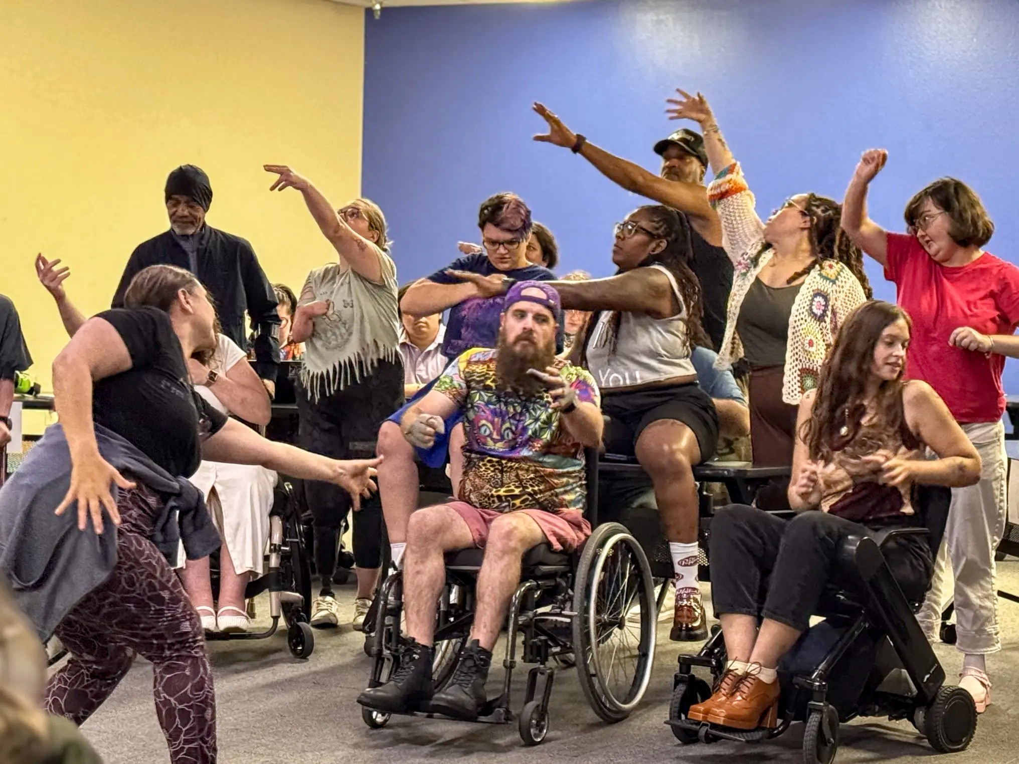 Black and white adult men and women rehearse a dance number in a bright room. Three use wheelchairs. Several reach their arms overhead.
