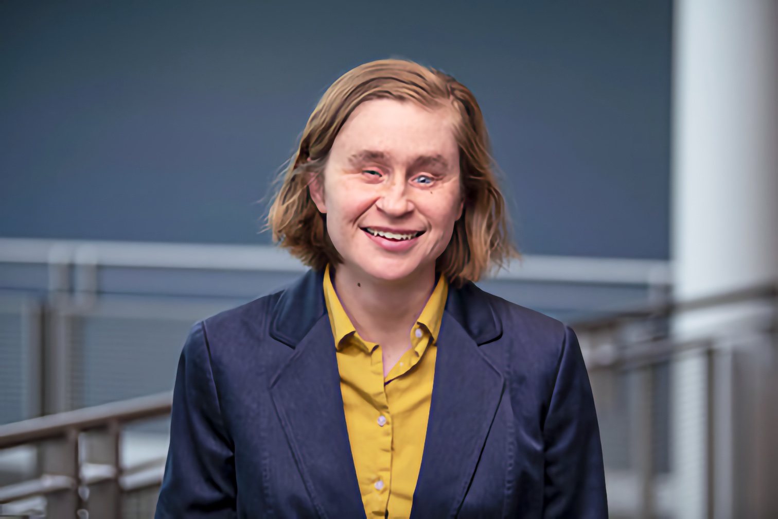A smiling middle-aged woman with medium-length light brown hair poses for a professional headshot in a well-lit indoor setting.