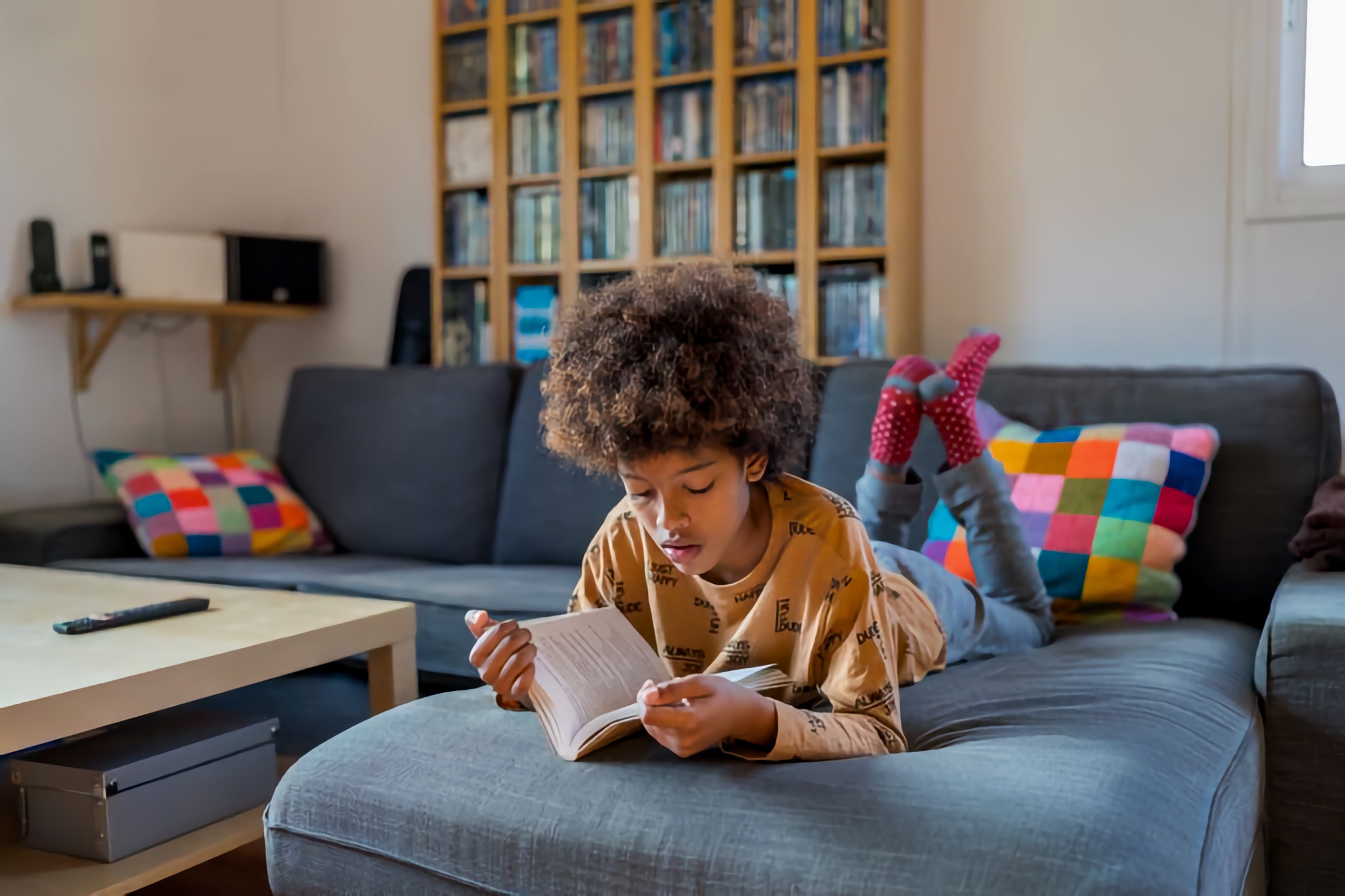 Brown-skinned later child lying on a gray sectional sofa reading a small paperback; head propped on hands, red-spotted socks up behind them; colorful patchwork pillows and a wall of shelving with books and media in the background; bright daytime living-room light.