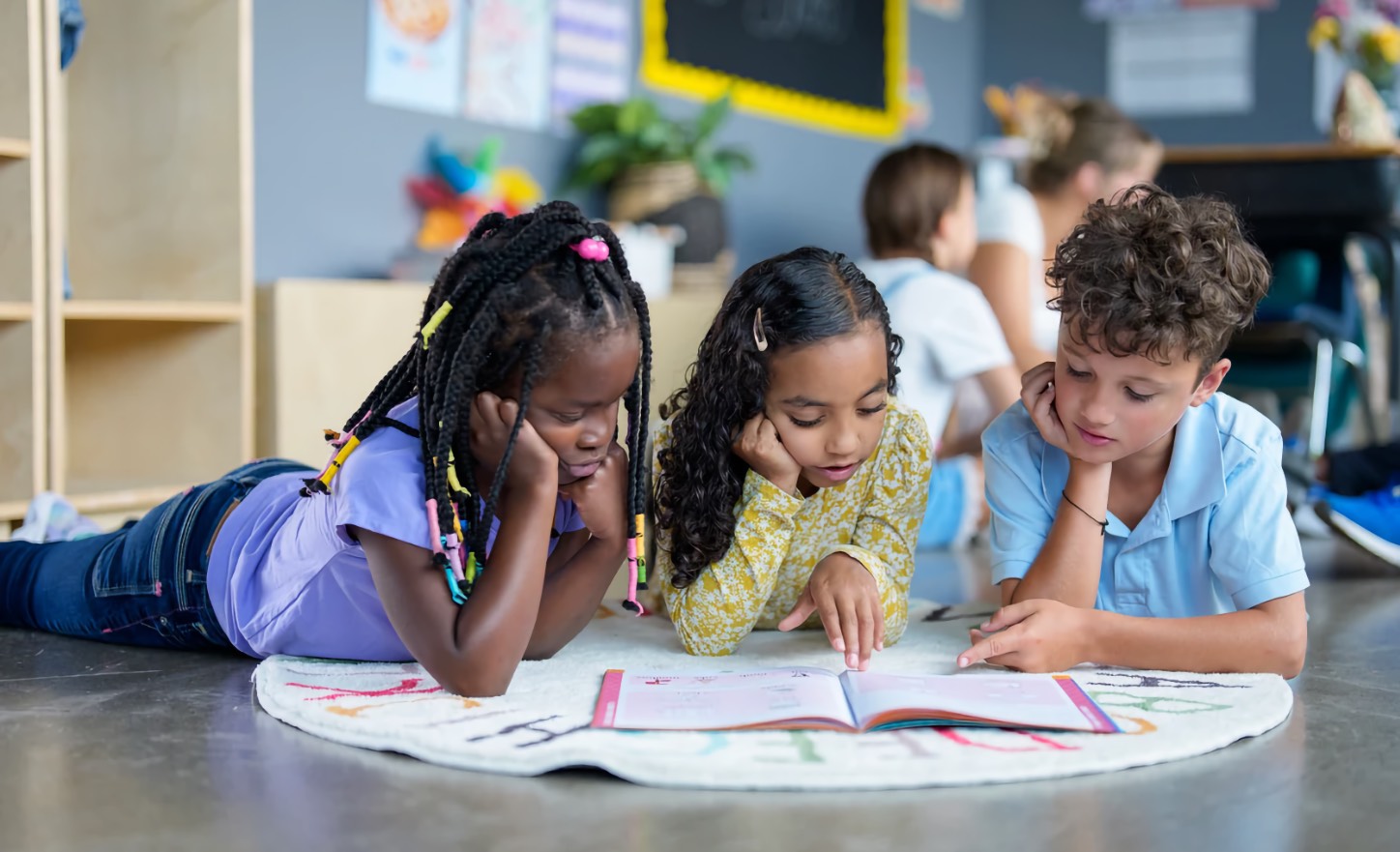 A Black girl, a girl, and a boy, all children, lie on a classroom rug and read an open book together.