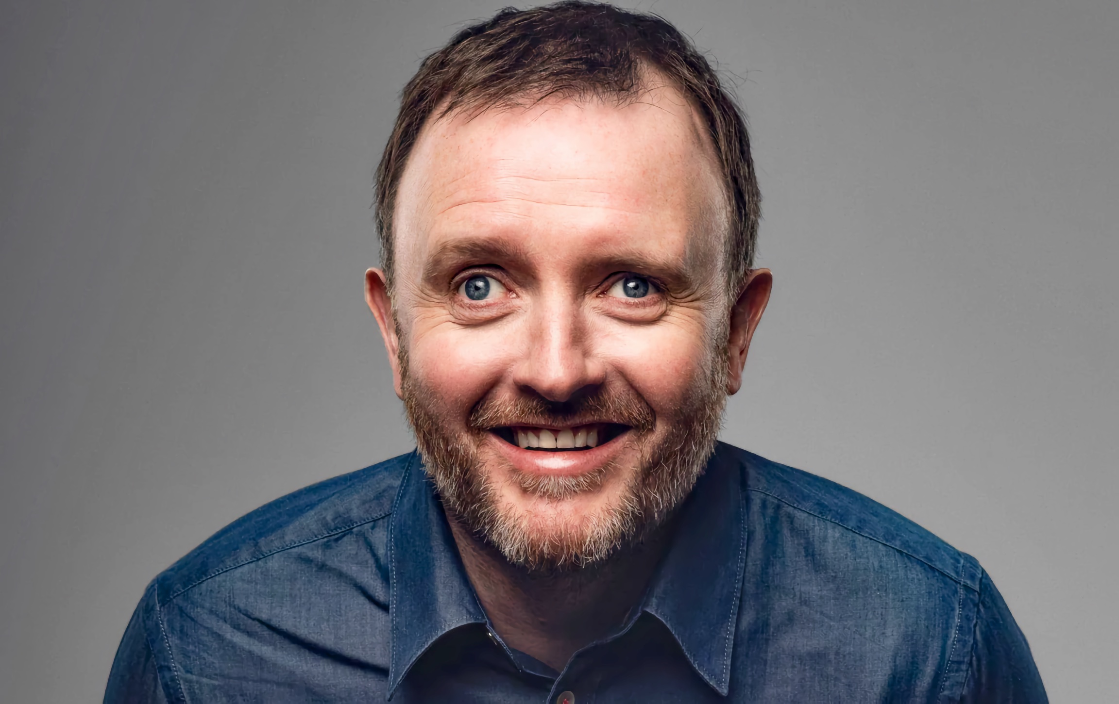 Middle-aged man smiles in a studio headshot; short brown hair, trimmed beard; denim shirt; neutral grey background.