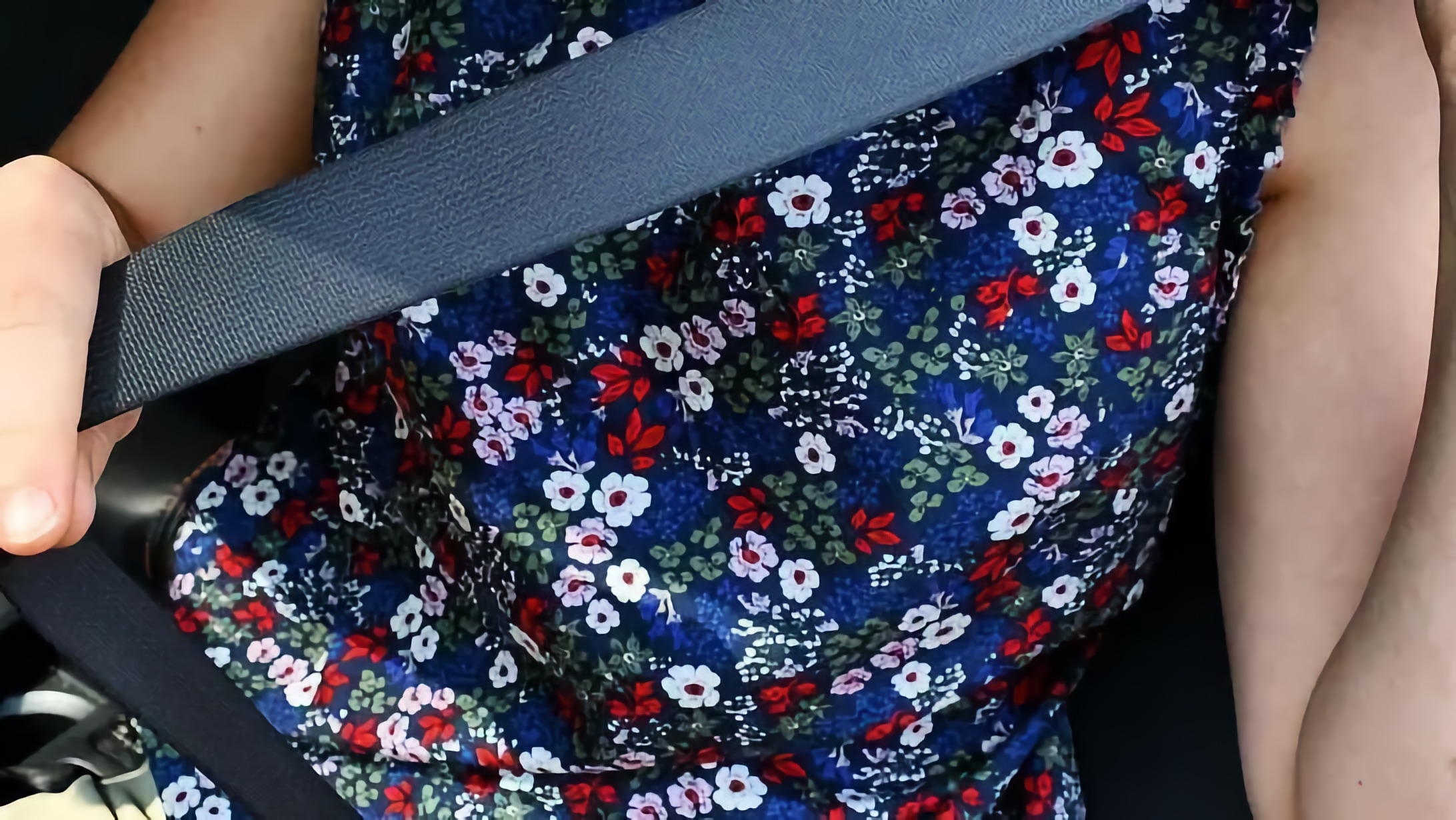 Hand pulls a dark seat belt across a blue flower-patterned top inside a vehicle