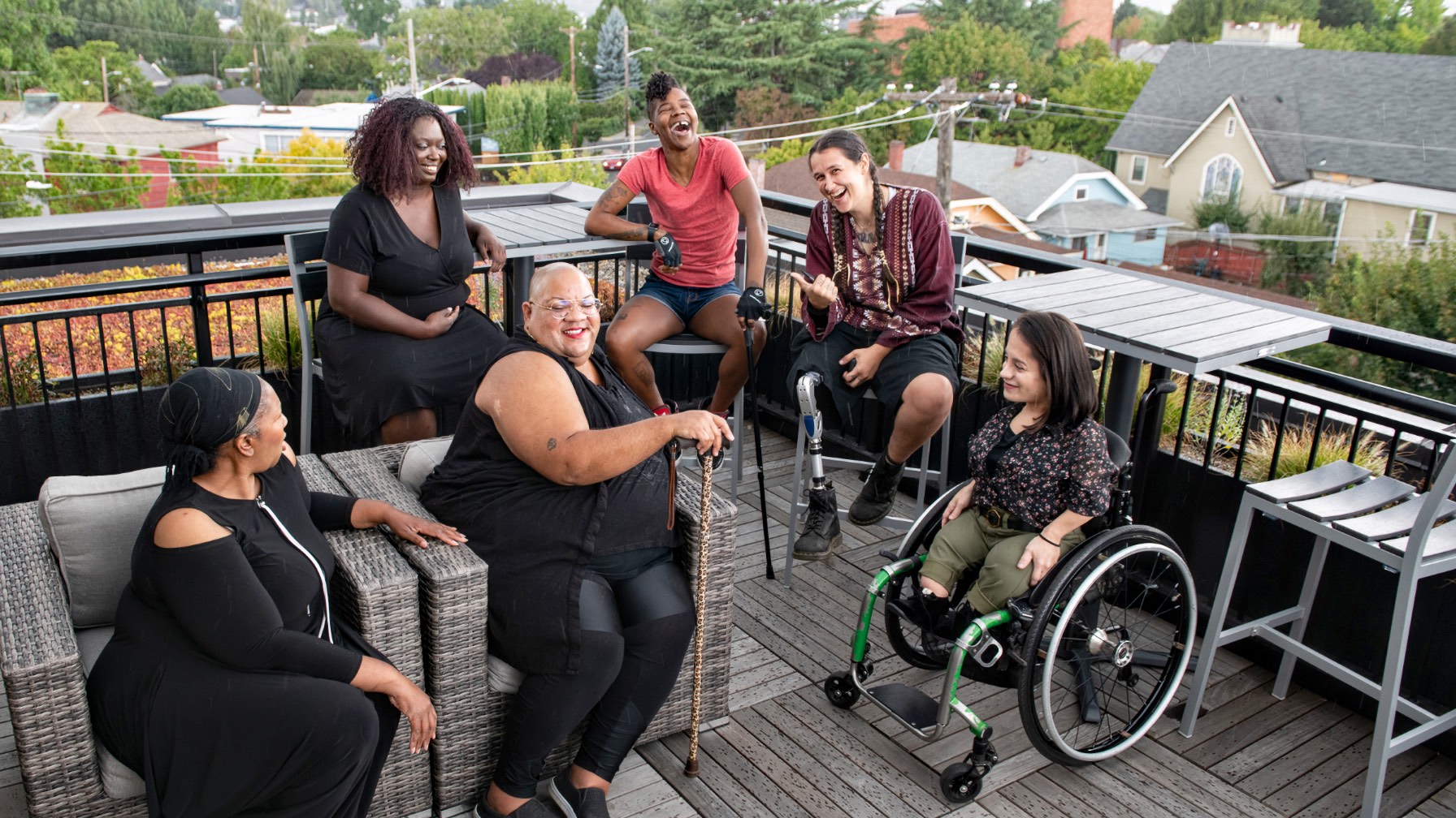 Six adults, mostly women, laughing on a rooftop deck. Four Black, two white. One uses a wheelchair, one a prosthetic leg, two use canes or crutches.