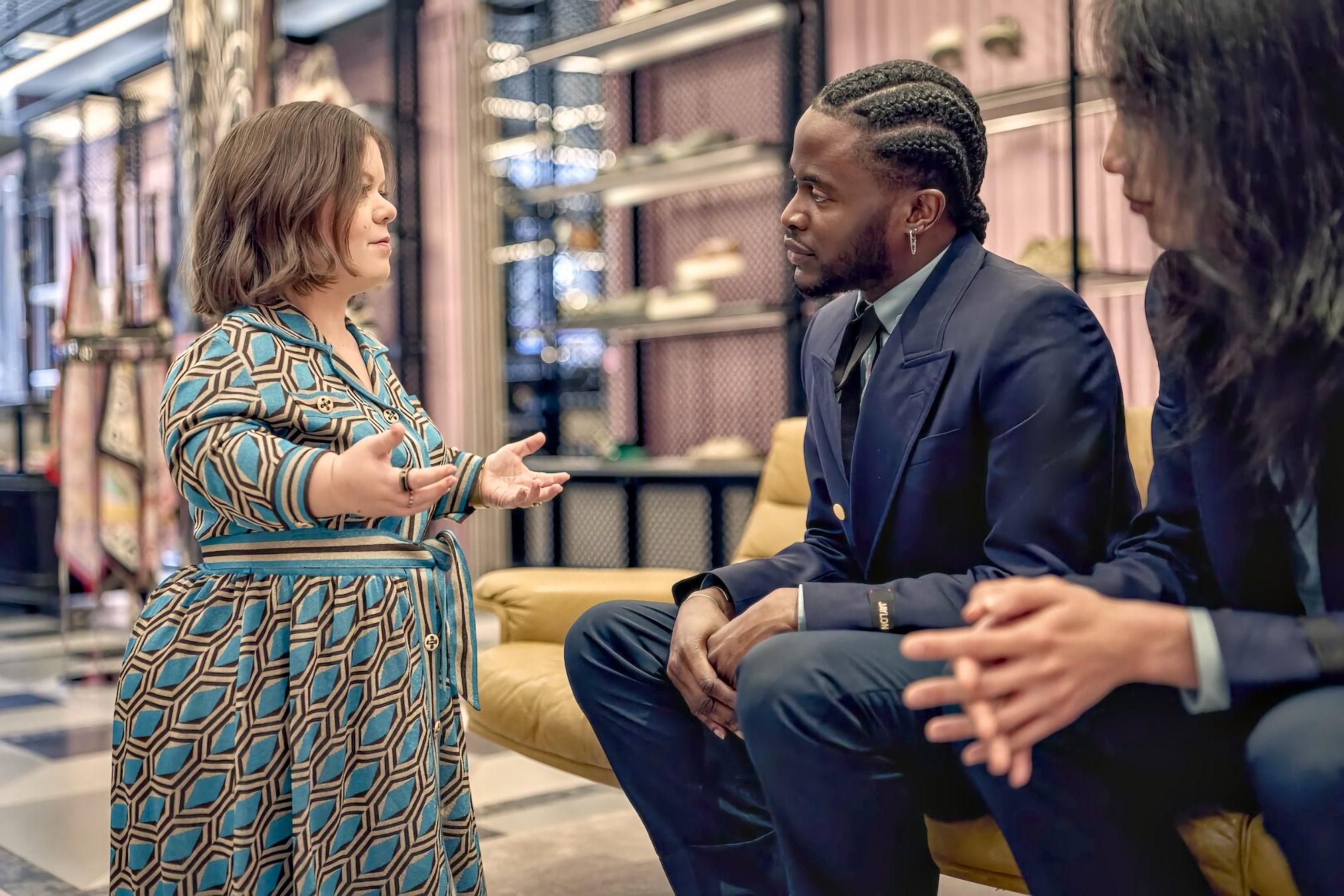 Woman in a blue geometric dress stands and gestures while speaking to two seated men in dark suits on a yellow sofa; shelves and merchandise in a stylish boutique are softly visible in the background.