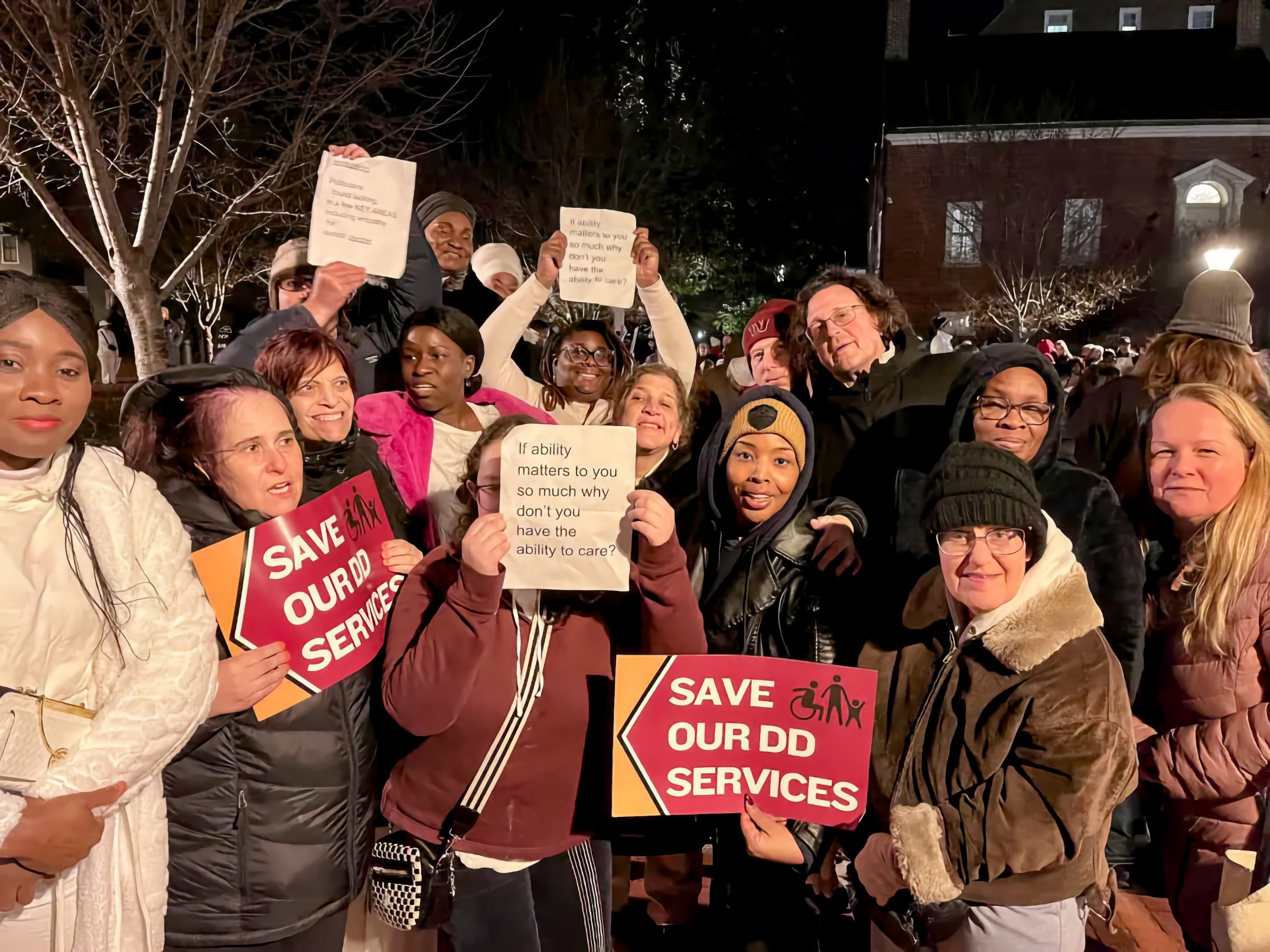 A multiracial crowd of approximately fifteen adults stands together outdoors at night. In the center foreground, a young Black woman wearing a maroon hoodie with white and black stripes holds a white handwritten sign reading "If ability matters to you so much why don't you have the ability to care?" To her left, a middle-aged white woman in a winter coat and glasses holds a similar handwritten sign with the same message. Throughout the crowd, multiple people hold bold orange-and-gold arrow-shaped signs reading "SAVE OUR DD SERVICES." Bare winter trees and a brick building are visible in the dark background.