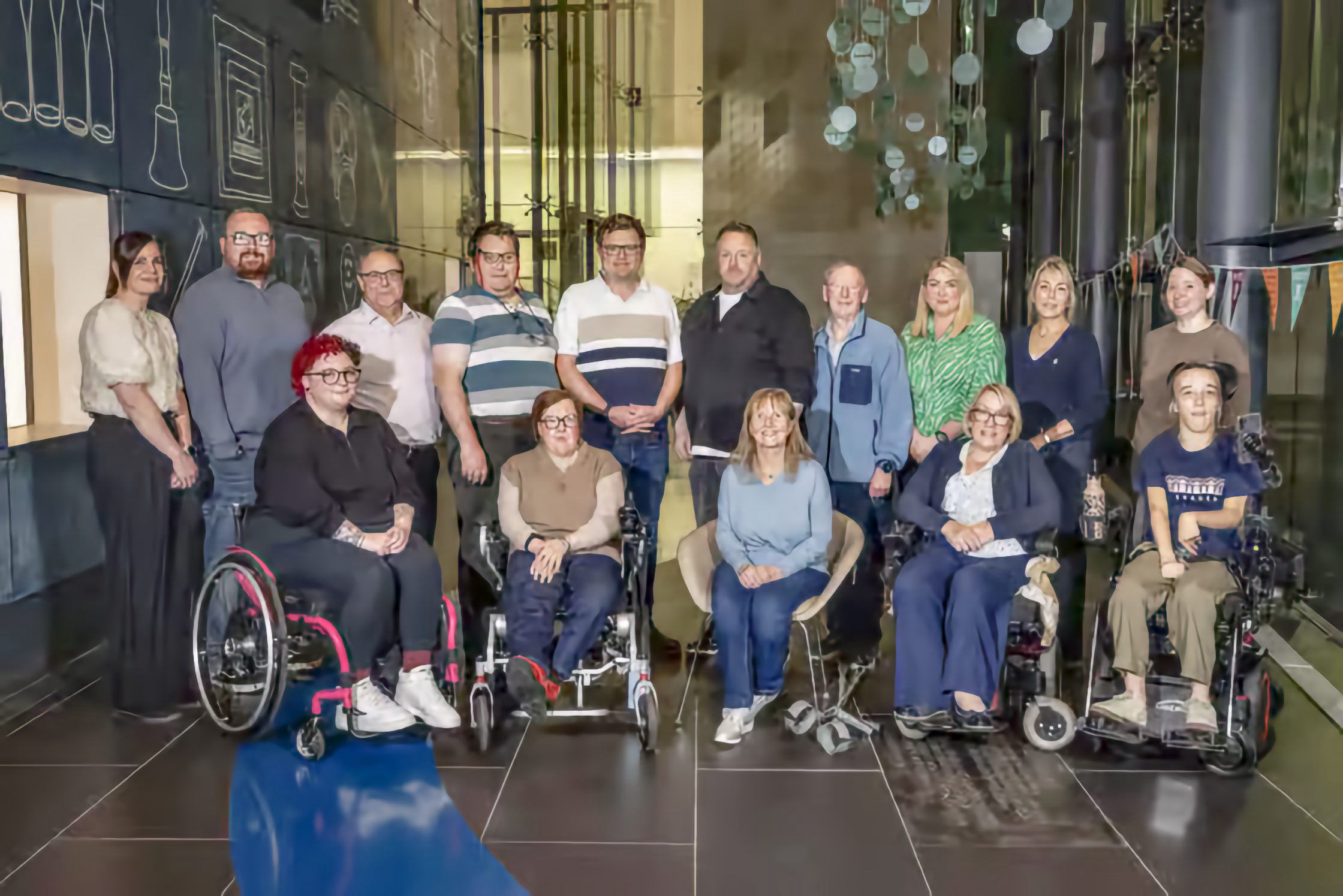 Men and women pose for a group photo indoors, some standing in rows and others seated in front, including four wheelchair users.