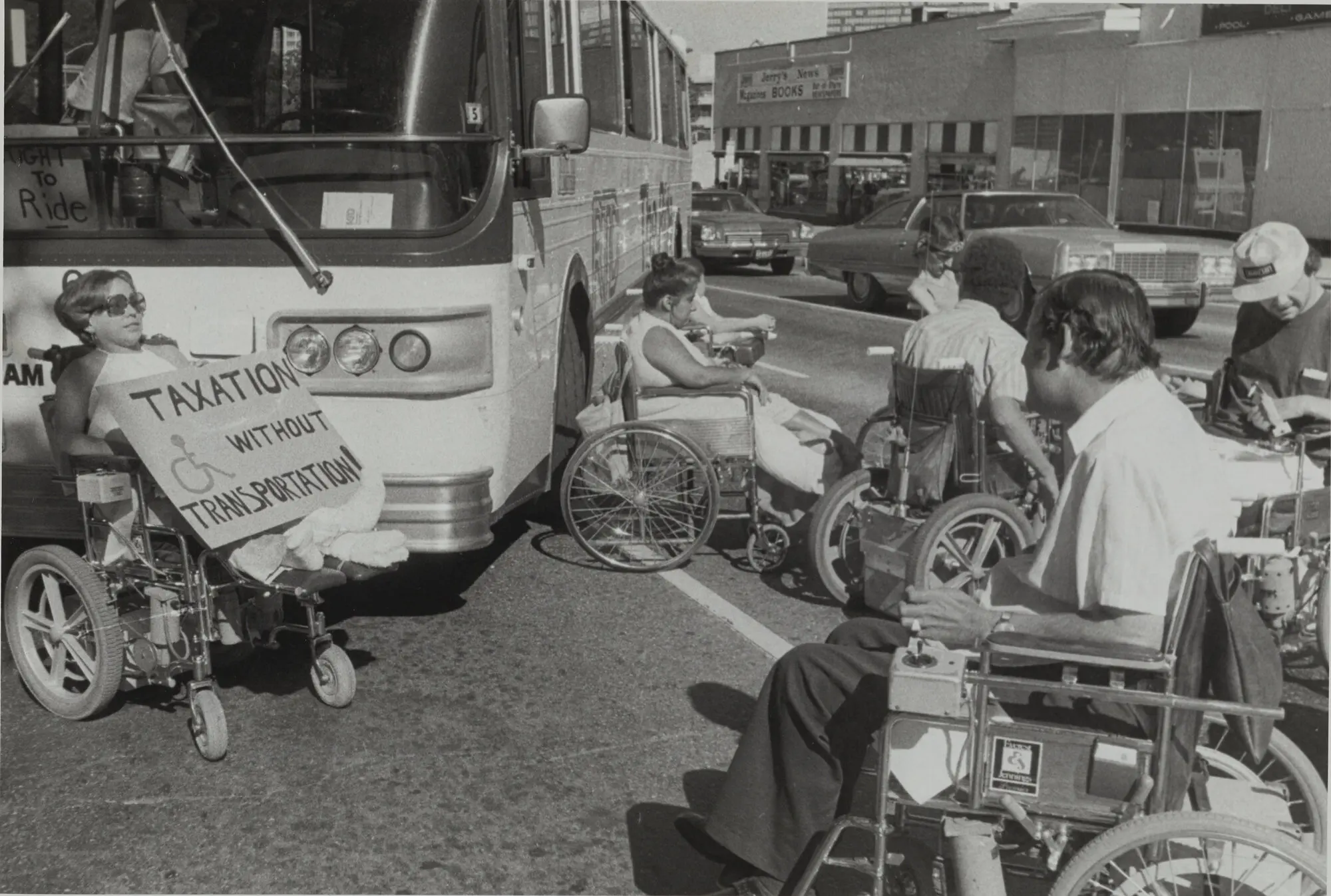 Several People using wheelchairs position themselves in front of and beside a large city bus on a busy street. One person holds a sign reading “Taxation Without Transportation” with a wheelchair access symbol, while cars wait behind.