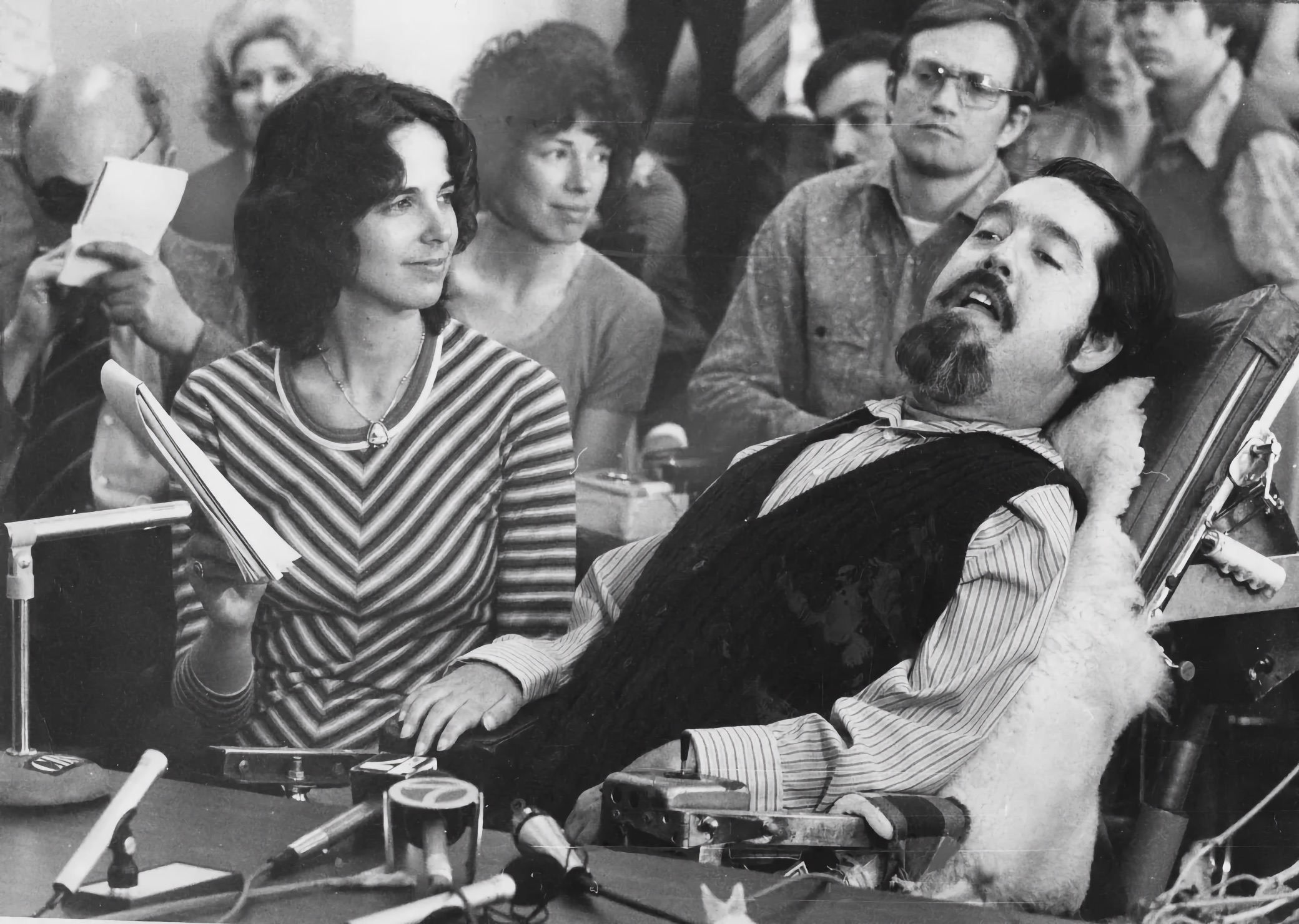 Ed Roberts, an adult man with dark hair, a mustache, and a goatee, sits at a hearing table in a powered wheelchair with a high back, headrest, and a gooseneck control near his face; multiple microphones and loose cables line the table; Catherine Dugan sits to his left holding a notepad; a tightly packed audience watches from close behind.