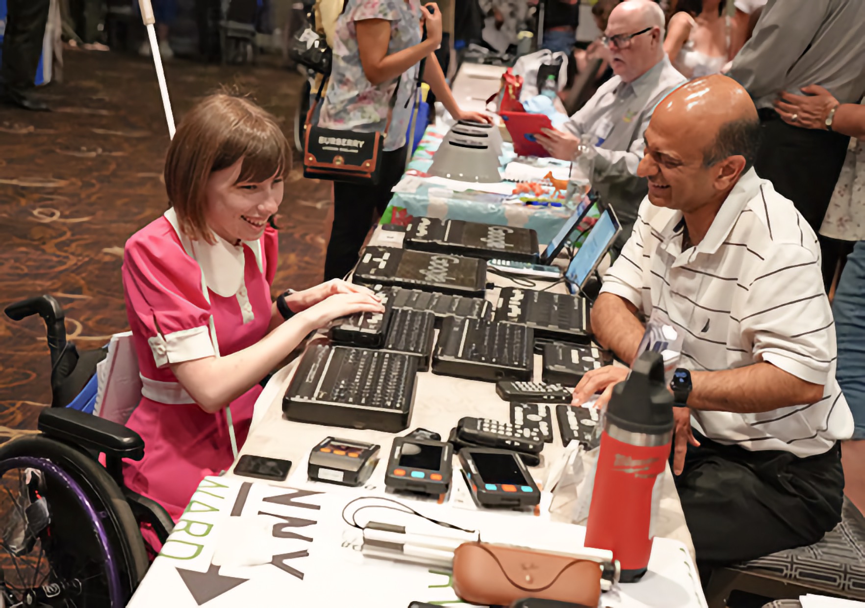 A Blind young adult woman seated in a manual wheelchair with colorful wheel accents reaches forward over an exhibit table and examines a Braille notetaker among an array of Braille devices and other access technology spread across the table surface. A man sits across from her, leaning forward and engaged in conversation. A crowded convention exhibit hall fills the background.