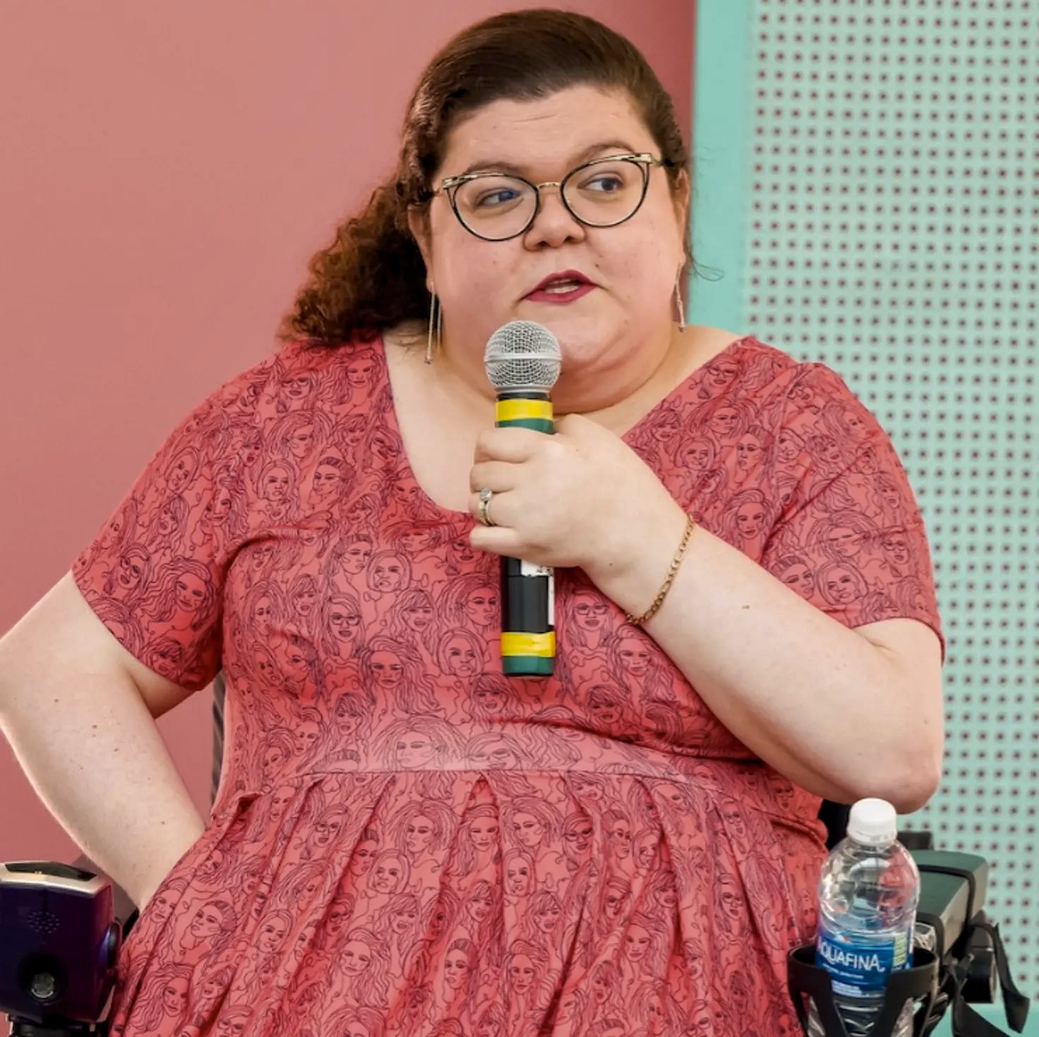 A woman seated in a power wheelchair speaks into a handheld microphone in front of a pink and teal wall.