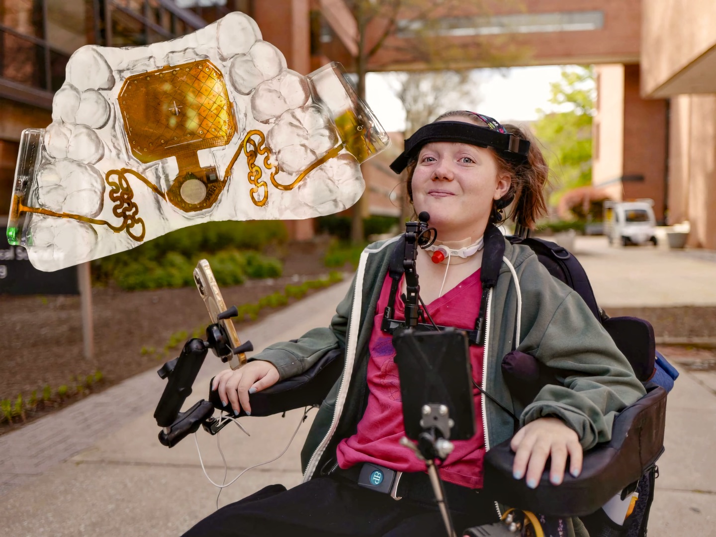 A teenage girl sits in a power wheelchair on a paved walkway beside a brick building, smiling toward the camera. She wears a black band across her forehead, a small device on a strap at her throat, and a tube at the base of her neck. A phone and a tablet are mounted on adjustable arms attached to her wheelchair. An enlarged view of the Mouthpad floats at the upper left, showing a clear molded dental retainer with an amber circuit panel and thin gold traces sealed inside it.