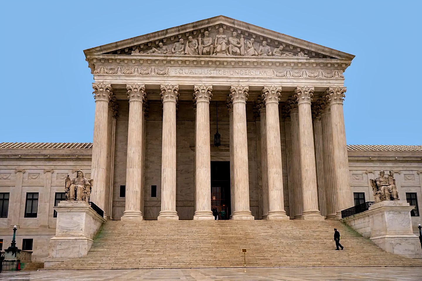 Exterior of the U.S. Supreme Court building in Washington, D.C. The white marble neoclassical facade features tall columns below a pediment with sculpted figures, and the words "EQUAL JUSTICE UNDER LAW" are carved into the frieze. A male security officer in a dark uniform stands on the lower right side of the broad marble staircase.