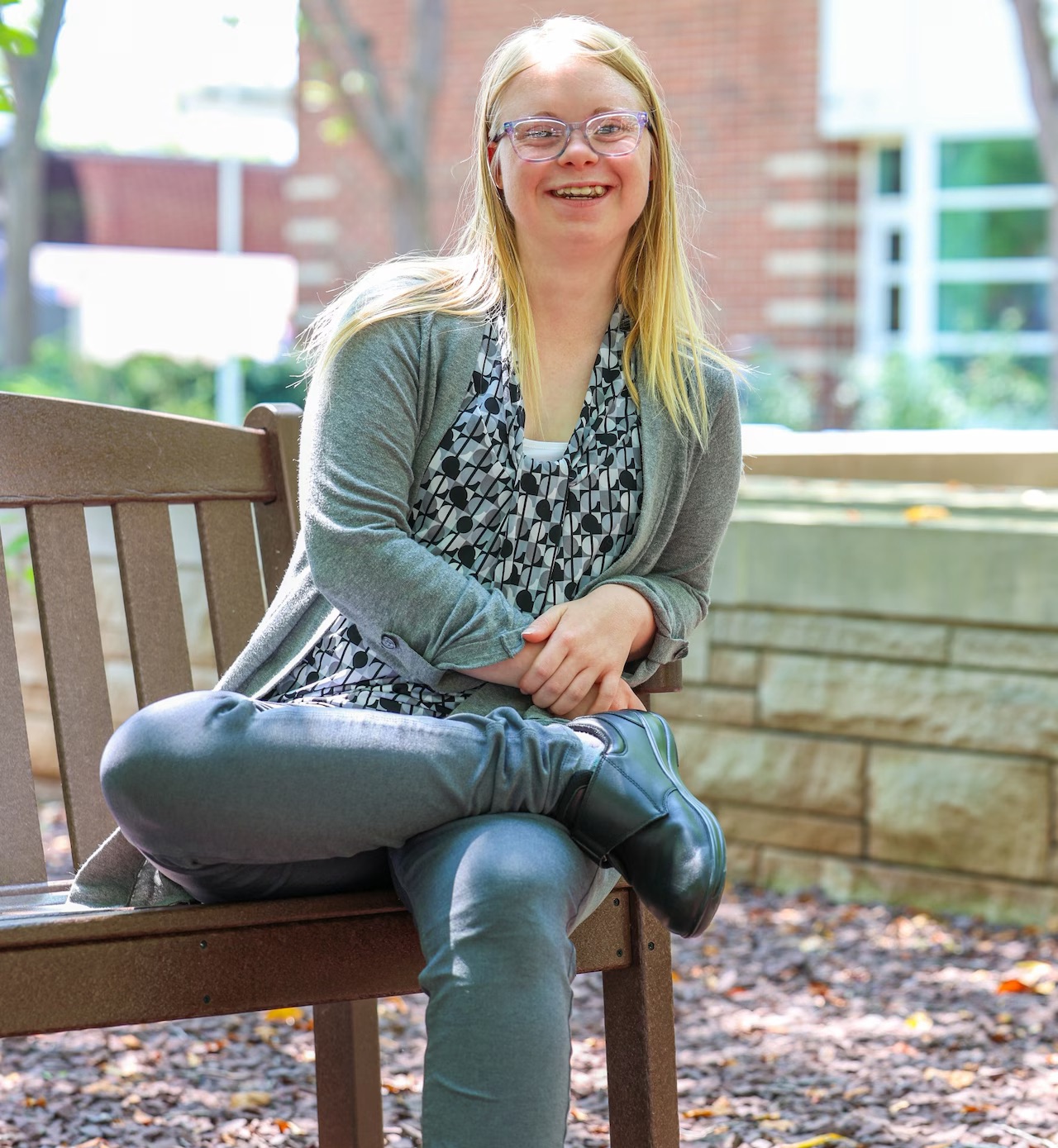 Sara Nye pictured with blond hair and glasses sits cross-legged on an outdoor bench, smiling in smart-casual gray attire.