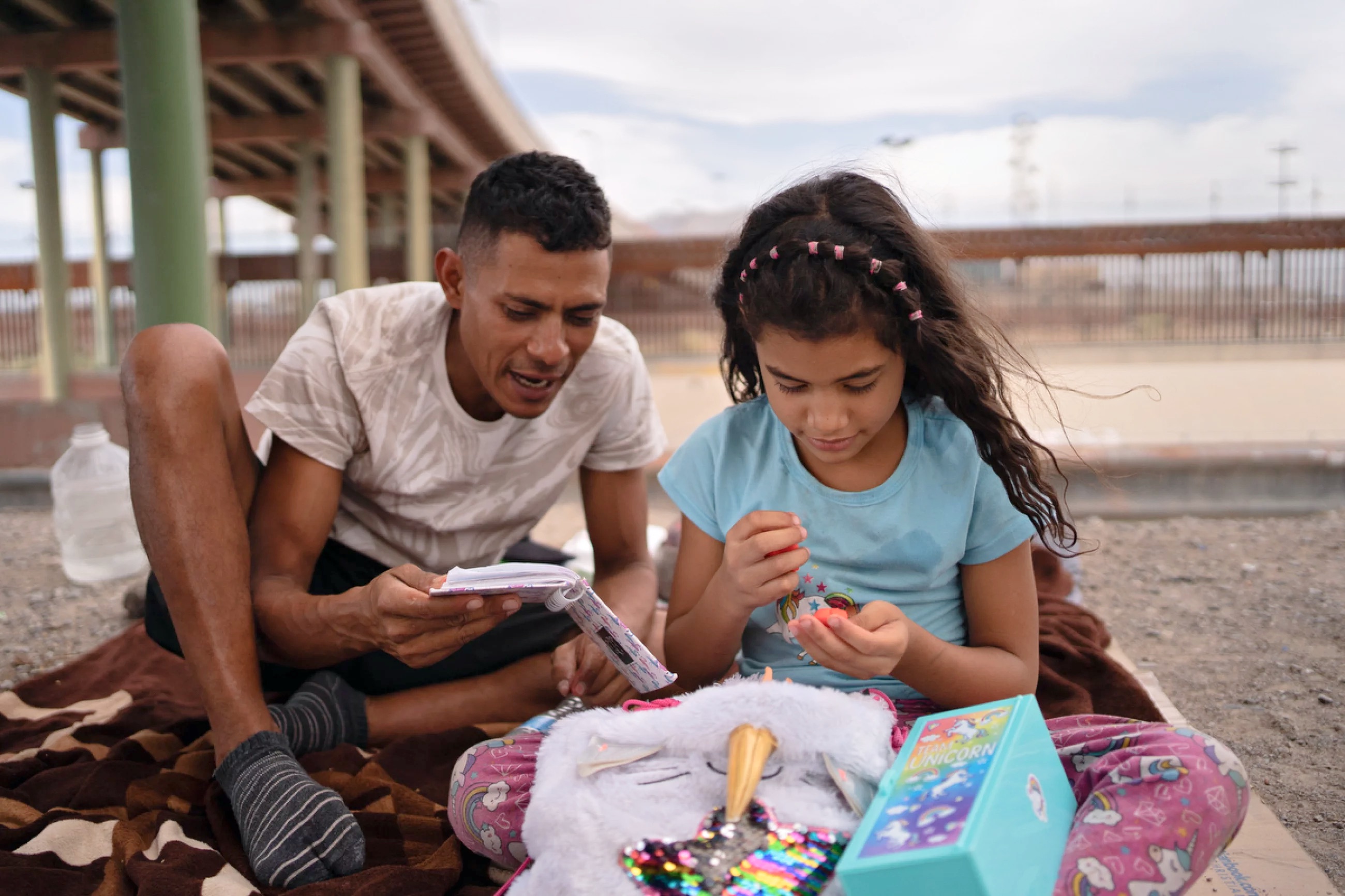 Man and girl sit on a blanket under a bridge; he reads a small notebook while she studies beads beside a unicorn backpack.