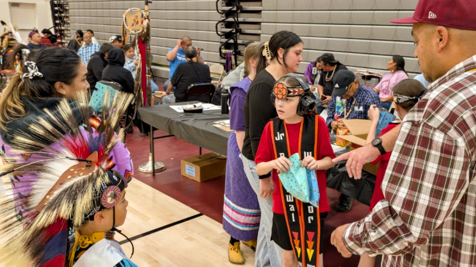 A Native American Autistic boy with headphones on holds a gift bag while a man points toward him and a dancer faces him.