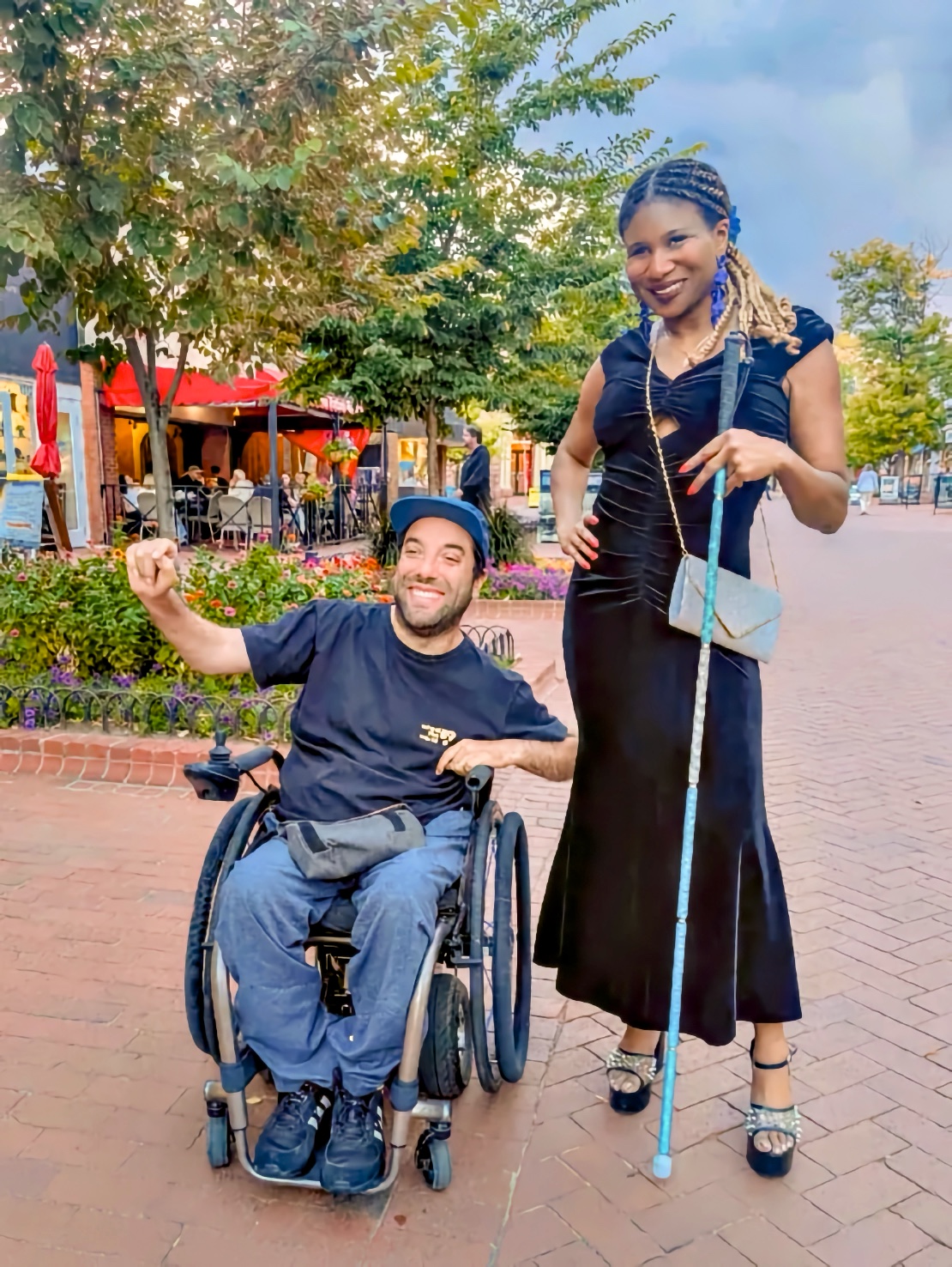 Adult blind Black woman standing on a brick pedestrian street with a bright blue bedazzled white cane; beside her, an adult man who uses a manual wheelchair smiles and lifts one arm; flowers, trees, and outdoor cafés in the background.