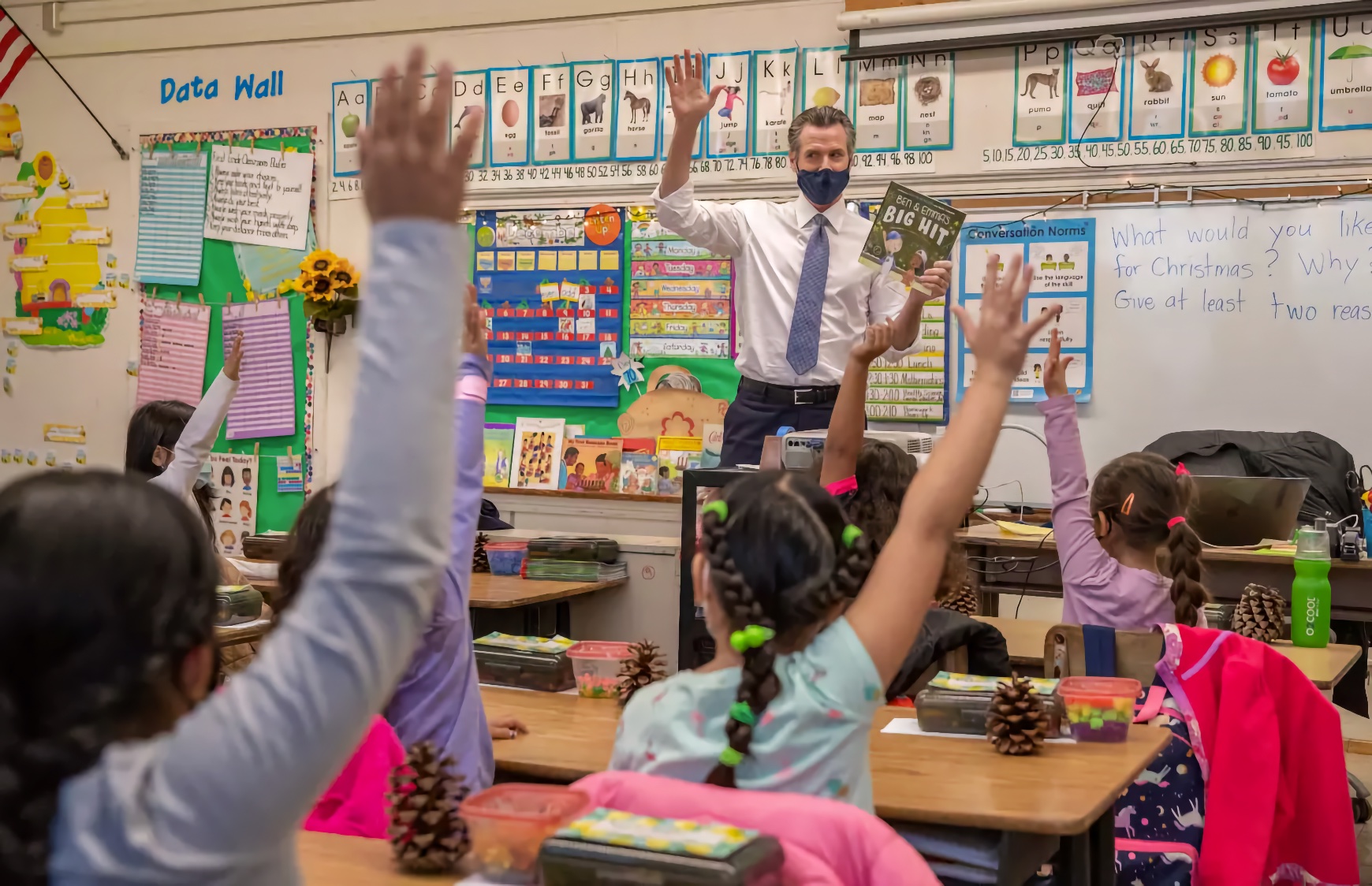 An adult male stands at the front of a colorful elementary classroom, holding up a children's book with both hands raised. Multiple young Latina girls seated at desks in the foreground eagerly raise their hands in response. The walls display an alphabet chart, a Data Wall bulletin board, and colorful educational materials.