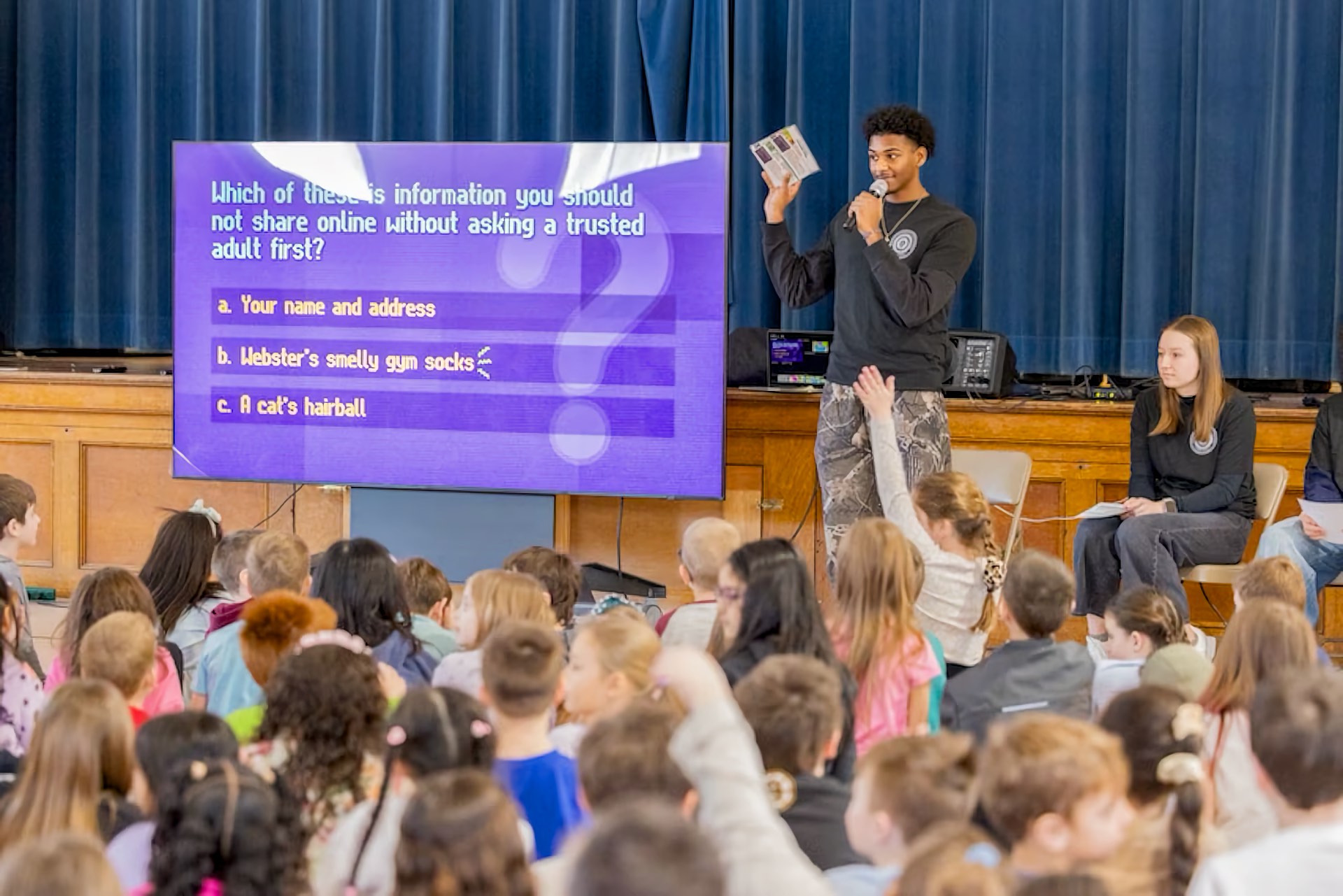 A Black male youth stands on a school auditorium stage holding a microphone in one hand and raising a booklet toward the audience with the other. A white female youth is seated on the stage behind him. A large group of children of mixed races and genders, ranging from young children to juniors, fill the auditorium seating with many hands raised. A large purple quiz slide is displayed on a screen to the left of the stage.