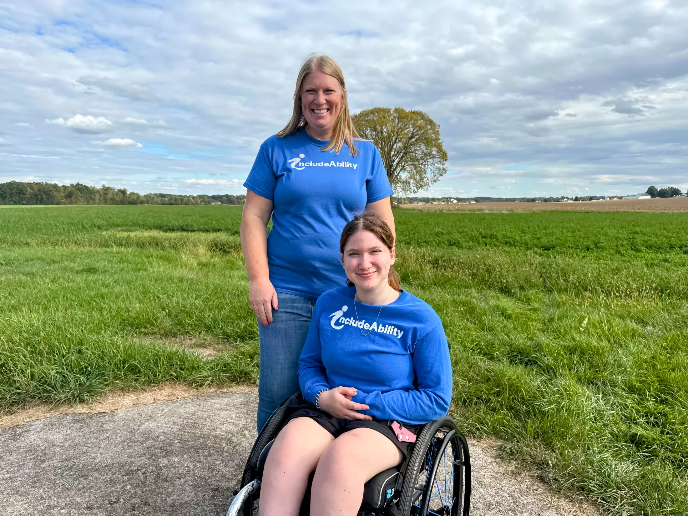 Young woman using a manual wheelchair sits on a paved path while an adult woman stands beside her; both wear blue “IncludeAbility” shirts; open farm fields and a cloudy sky stretch behind them.
