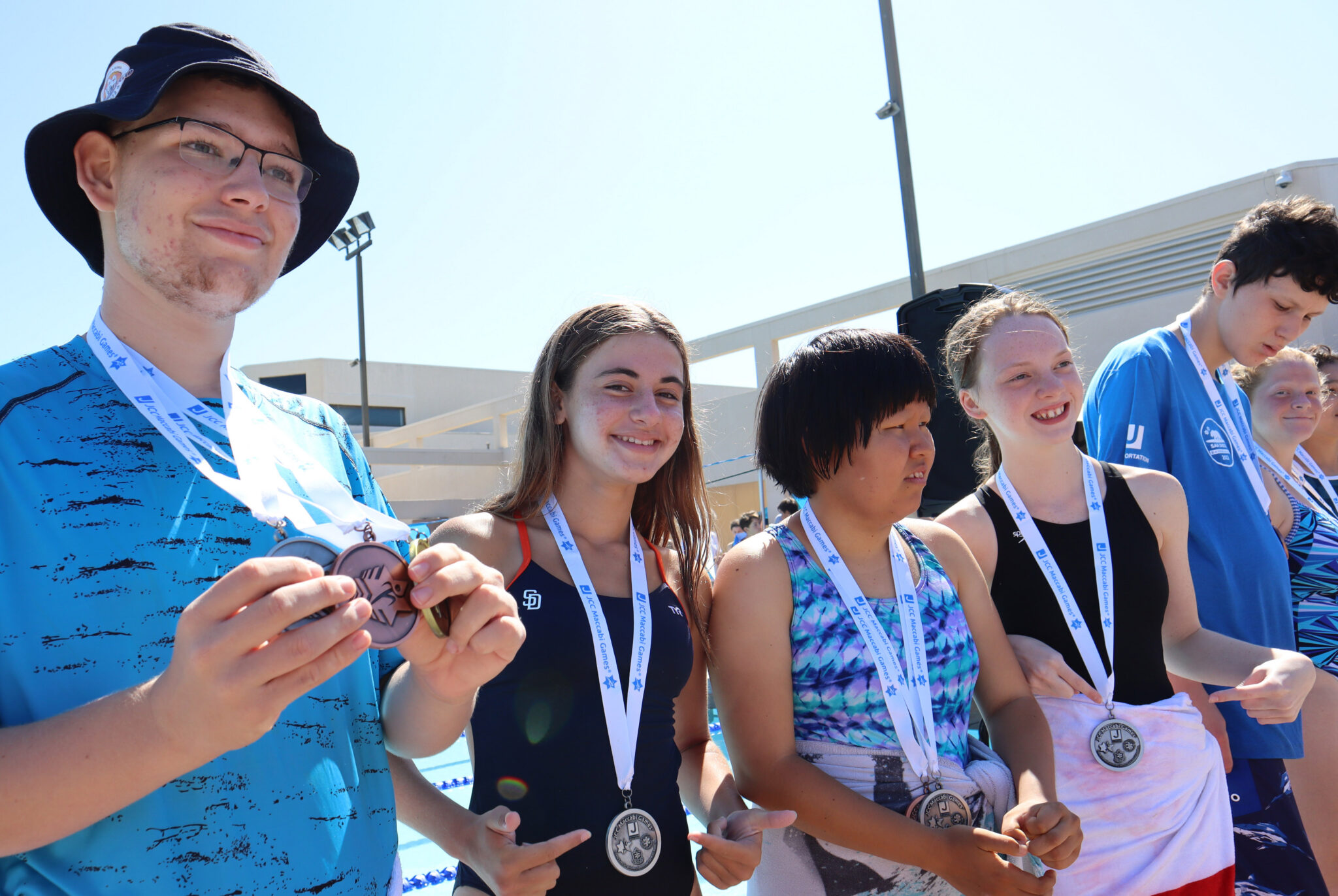 Outdoors beside a swimming pool, a white teen boy wearing glasses and a dark bucket hat holds up a bronze medal on a white ribbon while standing next to a line of teen girls and teen boys, including a white teen girl with long brown hair and an Asian teen girl with a short black bob; several youths wear medals and smile during an awards moment.