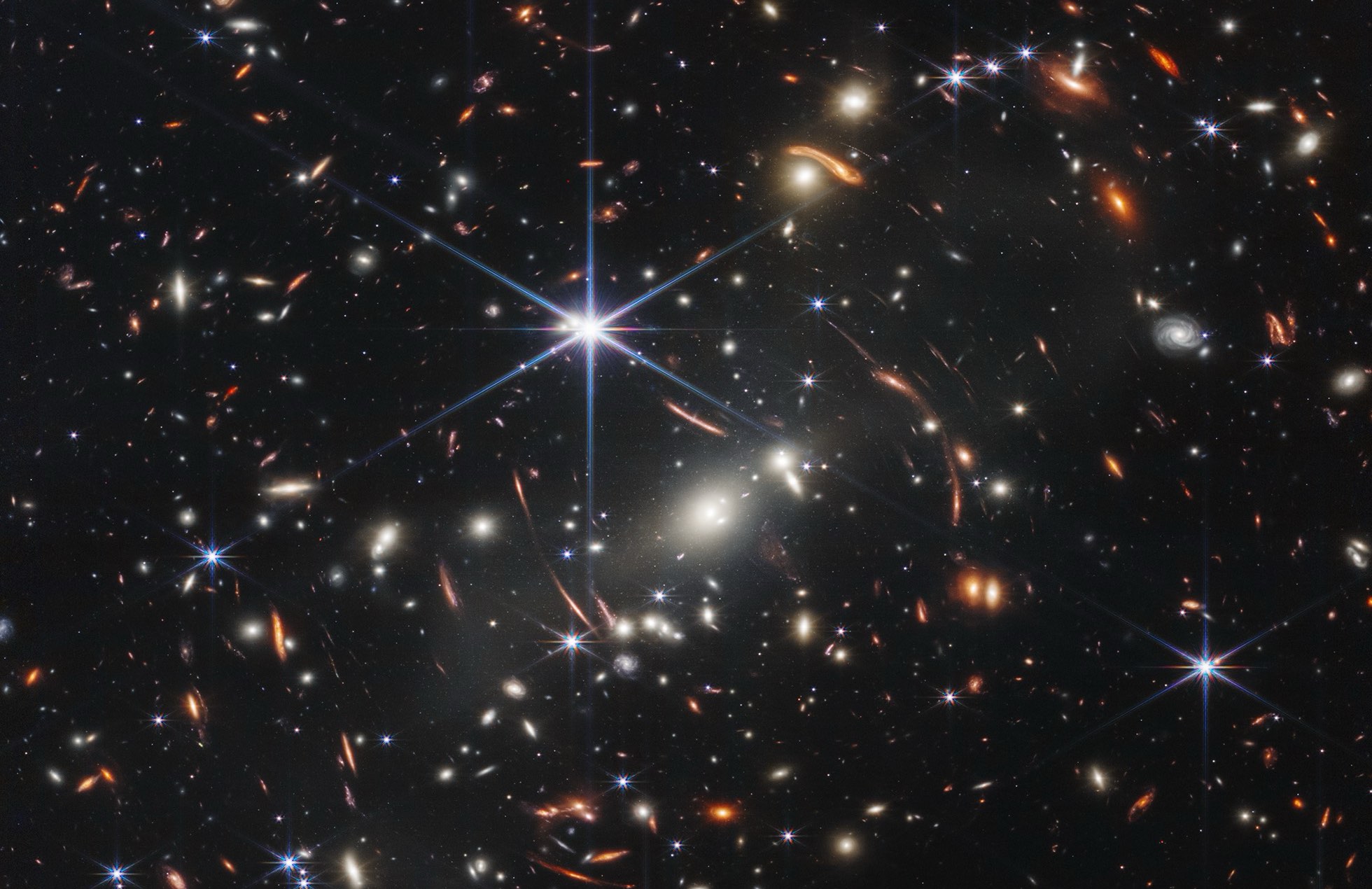 Thousands of small galaxies appear across this view. Their colors vary. Some are shades of orange, while others are white. Most appear as fuzzy ovals, but a few have distinct spiral arms. In front of the galaxies are several foreground stars. Most appear blue, and the bright stars have diffraction spikes, forming an eight-pointed star shape. There are also many thin, long, orange arcs that curve around the center of the image.