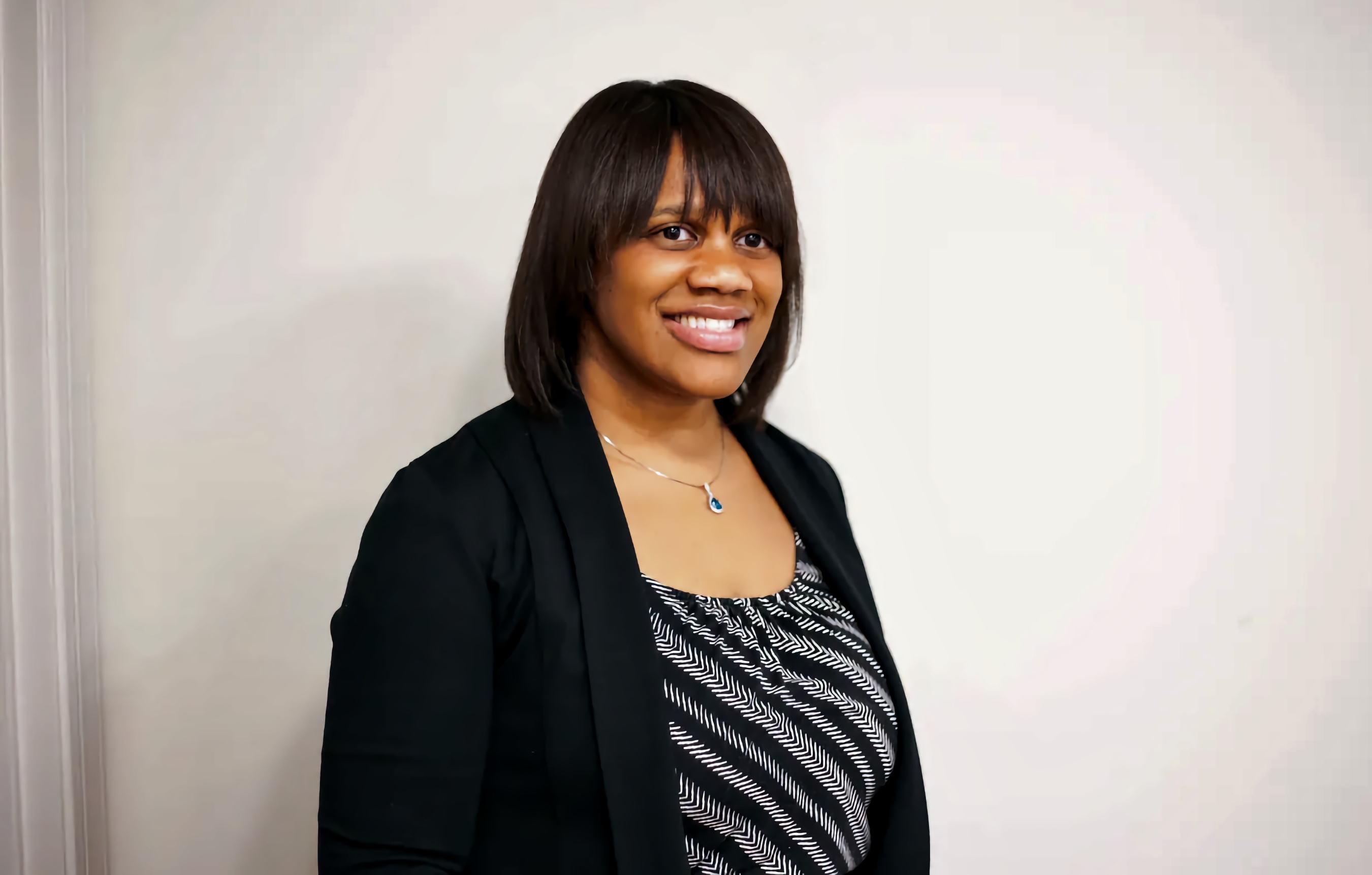 A Black woman with shoulder-length black hair and bangs is smiling in a professional headshot. She is wearing a black blazer over a black and white patterned blouse, with a silver necklace featuring a blue pendant. The background is a plain, off-white wall.