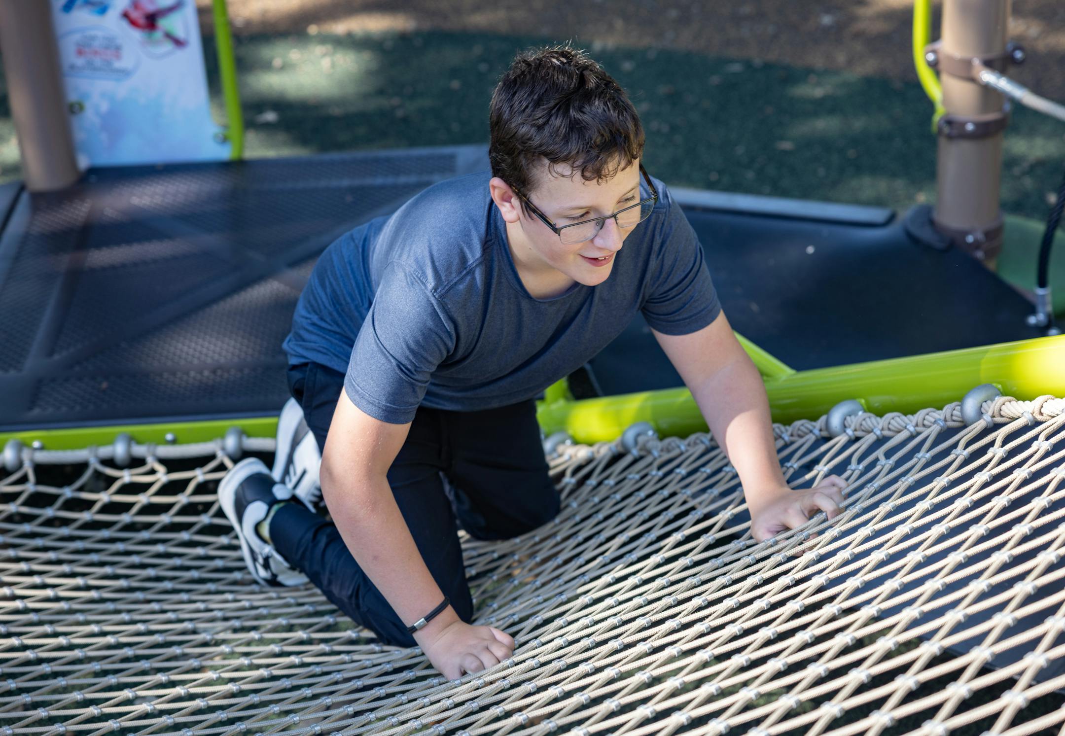 Teen boy climbs across a rope-net play surface on an inclusive playground.
